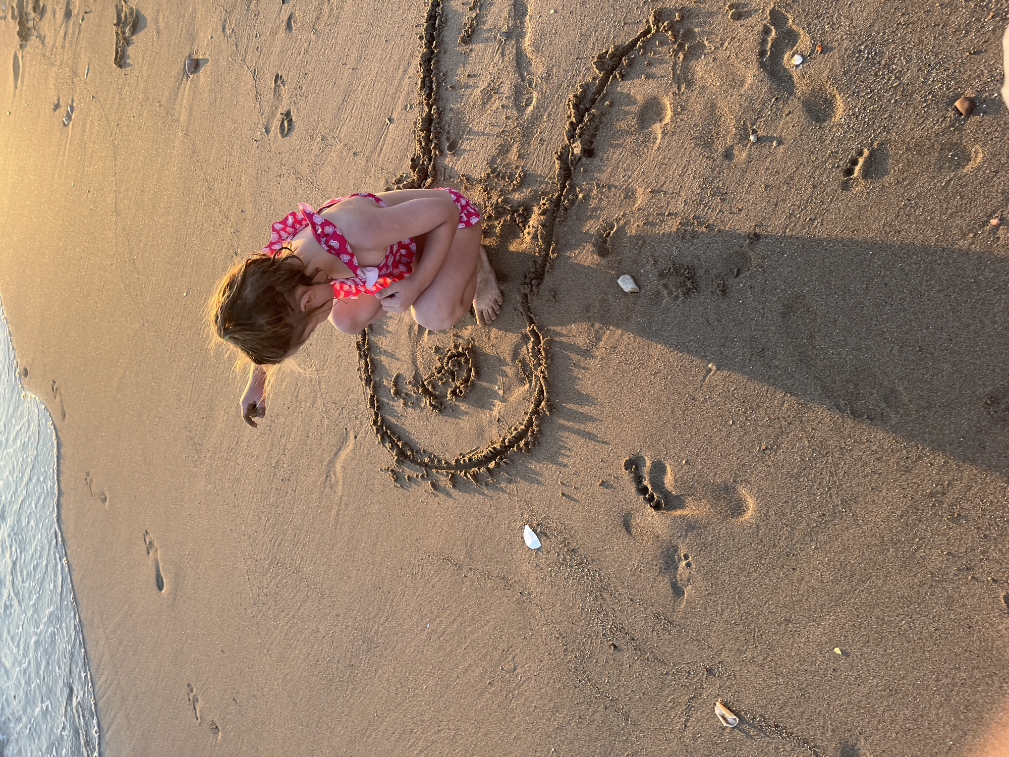 A young child, wearing a pink swimsuit with a seashell pattern, is crouched on a sandy beach at golden hour, drawing in the wet sand with their finger. The child's hair is wet and sandy. Footprints are visible in the sand, both from the child and others. The gentle waves of the sea are visible in the upper left corner of the frame. The scene is likely in Paleo Faliro, Greece, given the context.