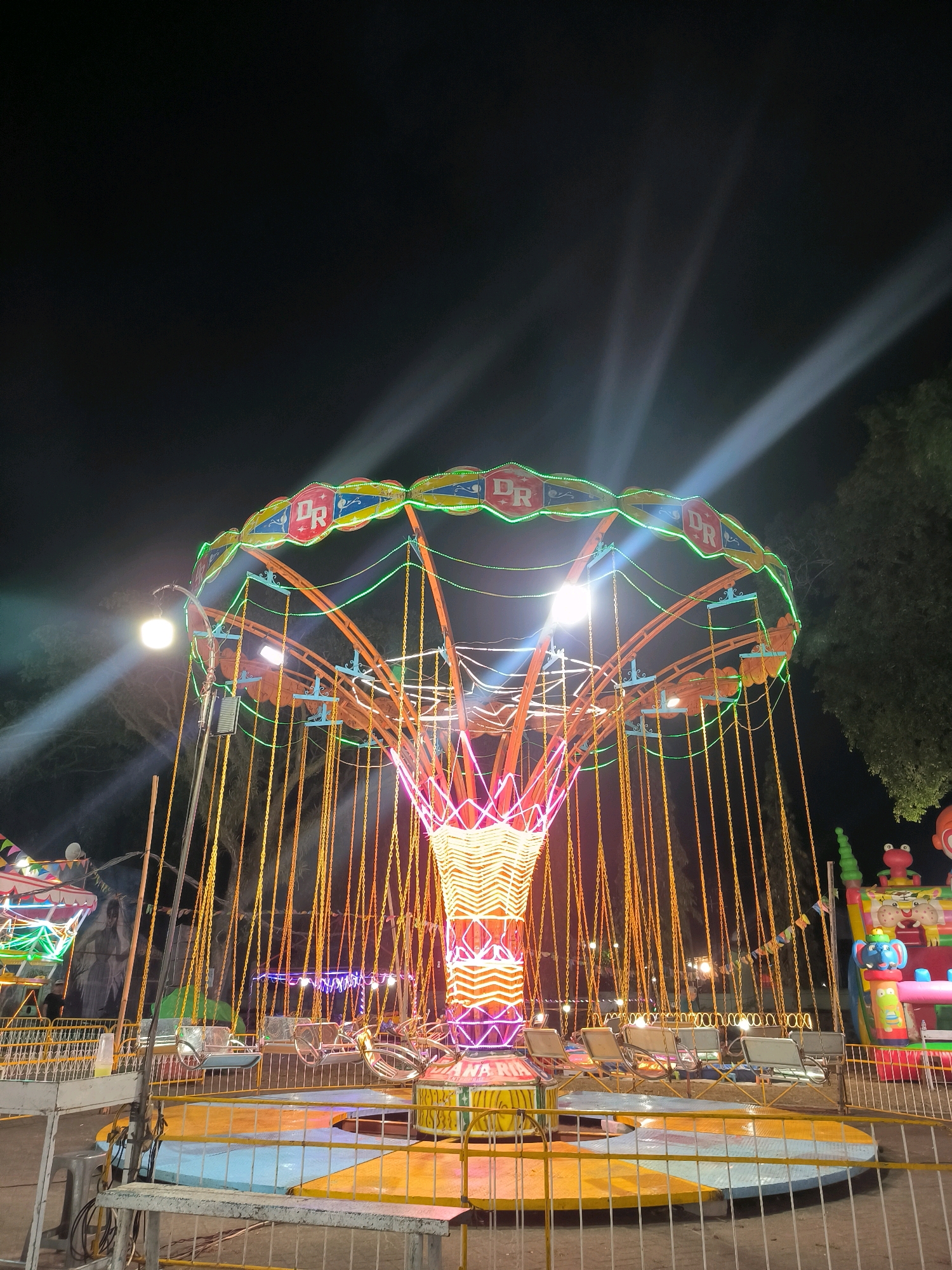 This is a low-angle, nighttime photograph of a brightly lit amusement park ride in Ambarawa, Indonesia. The main subject is a swing ride, characterized by its central tower adorned with vibrant neon lights in red, yellow, and green. Numerous chains hang from the circular top of the ride, each suspending a seat. The ride is surrounded by a safety barrier, and in the background, another inflatable attraction with cartoon characters is visible, along with trees and the dark night sky. Searchlights beam from the upper left of the frame. There are no people clearly visible in the image. The ground area around the ride is painted in blue and yellow. The overall scene suggests a festive atmosphere at an outdoor fair.