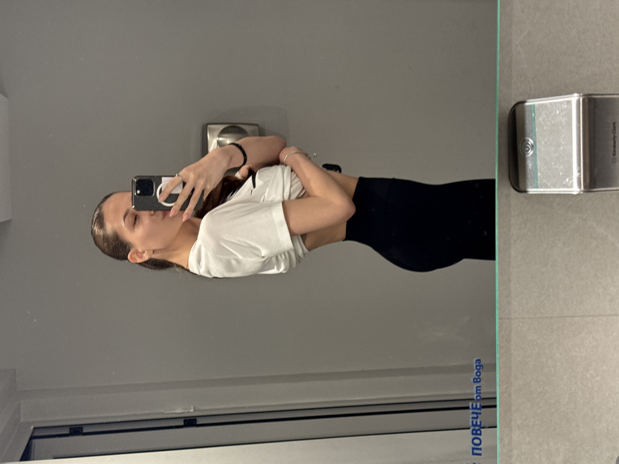 A mirror selfie captures a young woman indoors, likely in a public or commercial facility in Sofia, Bulgaria. The woman, with dark hair pulled back, is wearing a white short-sleeved top and black leggings. She holds a dark-colored smartphone with a light-colored circular grip in her right hand, positioned to take the photo. Her left arm is held across her midsection, and she looks down towards the phone.
The setting appears to be a modern indoor space, possibly a restroom or changing room. On the right side of the image, a wall-mounted dispenser is visible, which has faint text indicating "Kimberly-Clark". The wall and floor surfaces on the right are covered in light grey textured tiles. The large mirror reflects a plain grey wall and a white, window-like structure with dark horizontal lines at the bottom.
Near the bottom right corner of the mirror, blue text is visible, which reads "ПОВЕЧЕ om Вода". This translates from Bulgarian to "MORE than Water," reinforcing the Sofia, Bulgaria location context. The lighting is even, suggesting artificial indoor illumination. The overall impression is a casual, everyday moment captured through a mirror reflection.