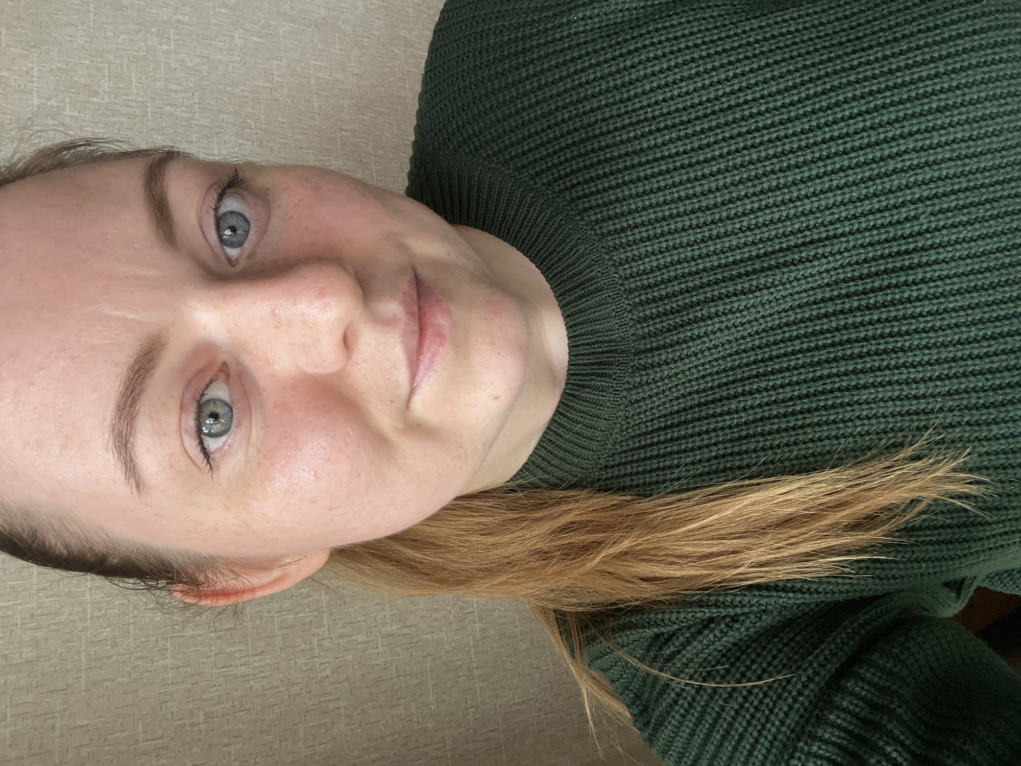 A close-up, slightly angled selfie shows a woman with fair skin and light brown hair. She has blue eyes and freckles on her face. She wears a dark green, knitted sweater with a high collar. The background is a light beige or grey textured surface, possibly a wall. No information suggests the photo was taken in Раздолье, Russia.