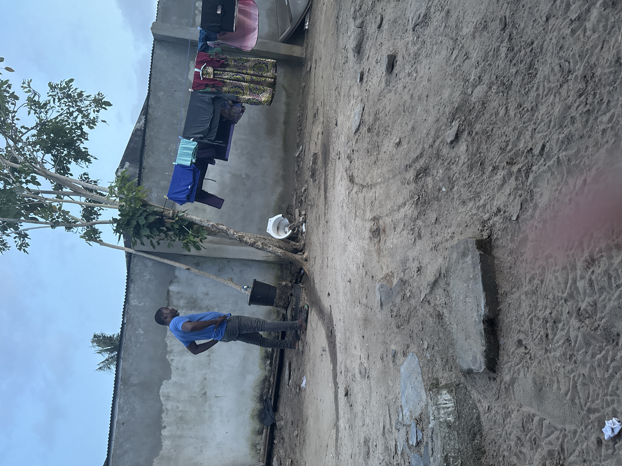 In Badagry, Nigeria, a man in a blue t-shirt and grey pants stands with his hands on his hips, facing away from the camera and looking towards a concrete wall. In the foreground, the ground is covered in dry earth and sand, with scattered stones and a few pieces of white paper. A black bucket sits on a tire near the man, beside a tree. Clothes are hanging on a line in front of the wall, including a colorful dress, a pink cloth, and other garments. A broken white toilet bowl is visible near the tree, and a sink is further to the right. The sky above is overcast.