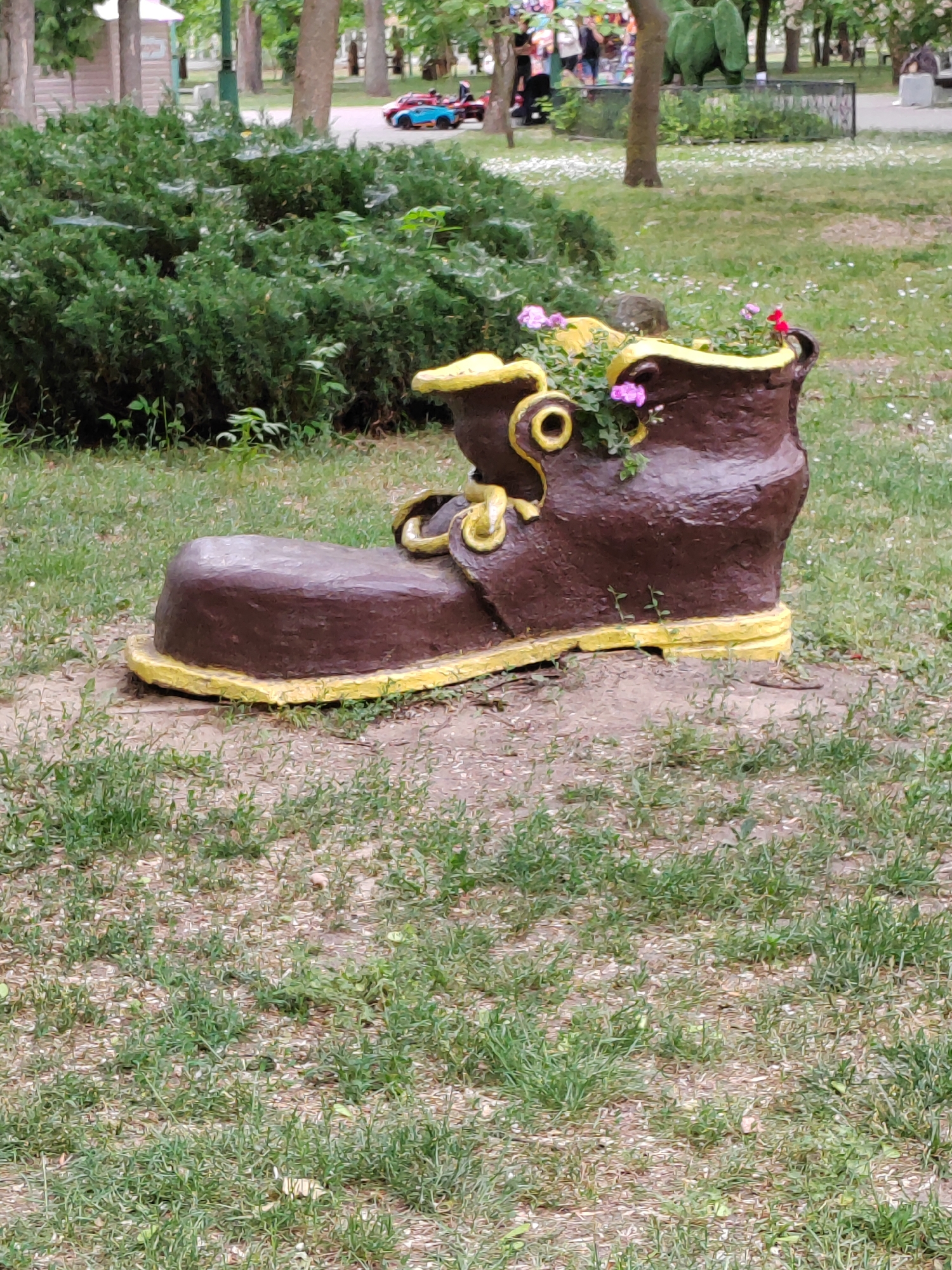 This image depicts a whimsical garden ornament shaped like a giant brown and yellow boot, situated outdoors on a grassy area. The boot serves as a planter, with small pink and red flowers growing from its opening. In the background, lush green bushes and trees are visible, along with a blurred park-like setting featuring a path, benches, and some indistinct figures. Further in the distance, there are children on a small rideable toy. The scene is bathed in natural daylight, suggesting it was taken during the daytime. There is no specific indication of the city of Brest, Belarus, in the visual information provided.