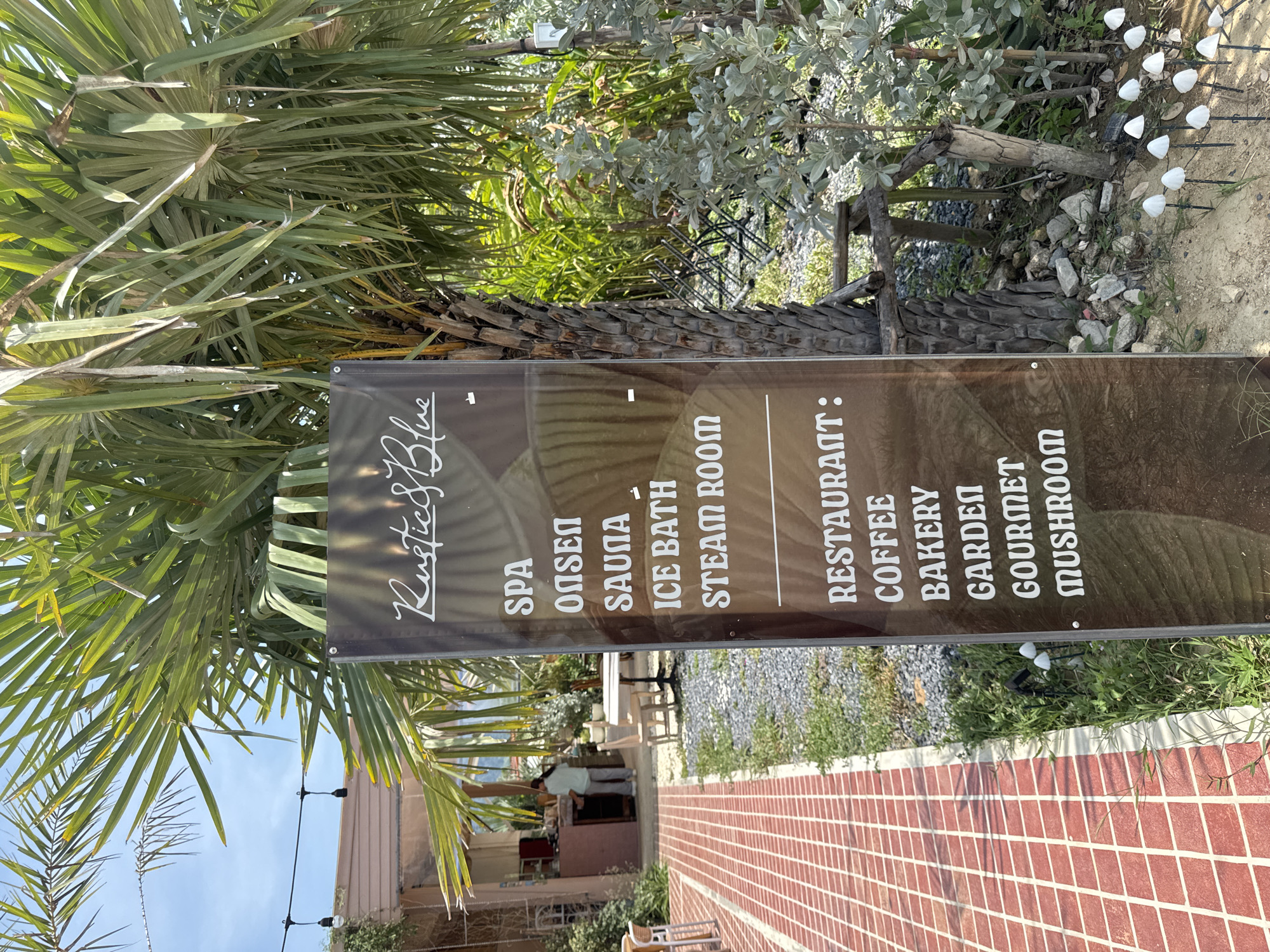 Here is a description of the image:
The image is a vertical shot of a sign for "Rustics Blue", with the sign listing the services offered, partially obscured by foliage. The services listed include spa, onsen, sauna, ice bath, steam room, restaurant, coffee, bakery, garden, gourmet, and mushroom. The sign is positioned beside a red brick walkway, bordered by greenery, and the background shows a building partially hidden by palm trees, beneath a clear blue sky. The location is likely Thep Krasatti, Thailand, given the query context. The photograph is taken at eye level, with the focus on the sign and the surrounding environment.