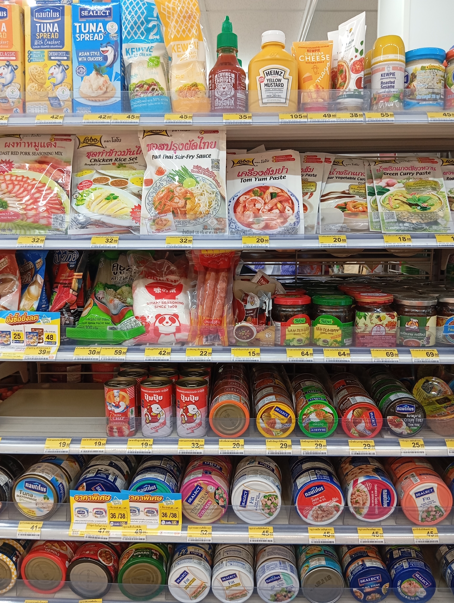 The image shows a shelf display of various food products in a retail setting, likely a convenience store or supermarket, located in Karon, Thailand. The products are neatly arranged on multiple shelves. The top shelf features an assortment of sauces and condiments, including sriracha, mustard, cheese spread, and roasted sesame paste. Brands like Sriracha, Heinz, Kewpie, and Sealeclt are visible.
The middle shelf is dedicated to various pastes and seasoning mixes, such as roast pork seasoning, chicken rice set seasoning, pad thai stir-fry sauce, tom yum paste, and green curry paste. Packaging includes brands like Lobo and Yum Yum. Also on this shelf are bags of what appears to be seasoning powder and packaged noodles. Canned goods are present on the lower shelves, including sardines and tuna. Brands like Nautilus, Sealeclt, and Lobo are identifiable. Prices are displayed below many of the products, with denominations in Thai Baht (฿). The lighting suggests an indoor retail environment. No people are visible in the frame.