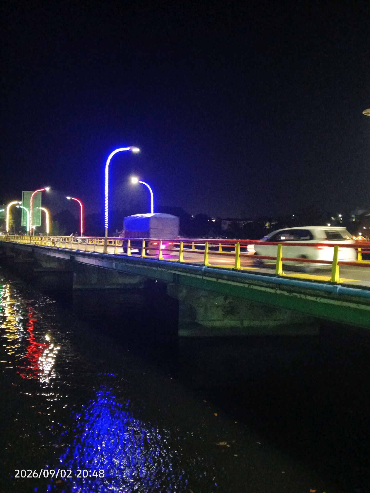 A night-time, long-exposure photograph captures a bridge adorned with colorful lights over a body of water. The bridge features yellow and blue railings, with green-painted supports visible underneath. Several streetlights with blue, red, and yellow illumination line the bridge, reflecting onto the dark water below in vibrant streaks of light. A white car is seen in motion on the bridge, blurred due to the long exposure. In the background, the silhouette of trees and buildings is dimly visible against the night sky. A large truck with a blue tarp covering its cargo is also present on the bridge. The date and time stamp "2026/09/02 20:48" is visible in the lower-left corner of the image. This scene is located in Pare-Pare, Indonesia, likely on the Pare-Pare Bridge, a prominent landmark in the city.