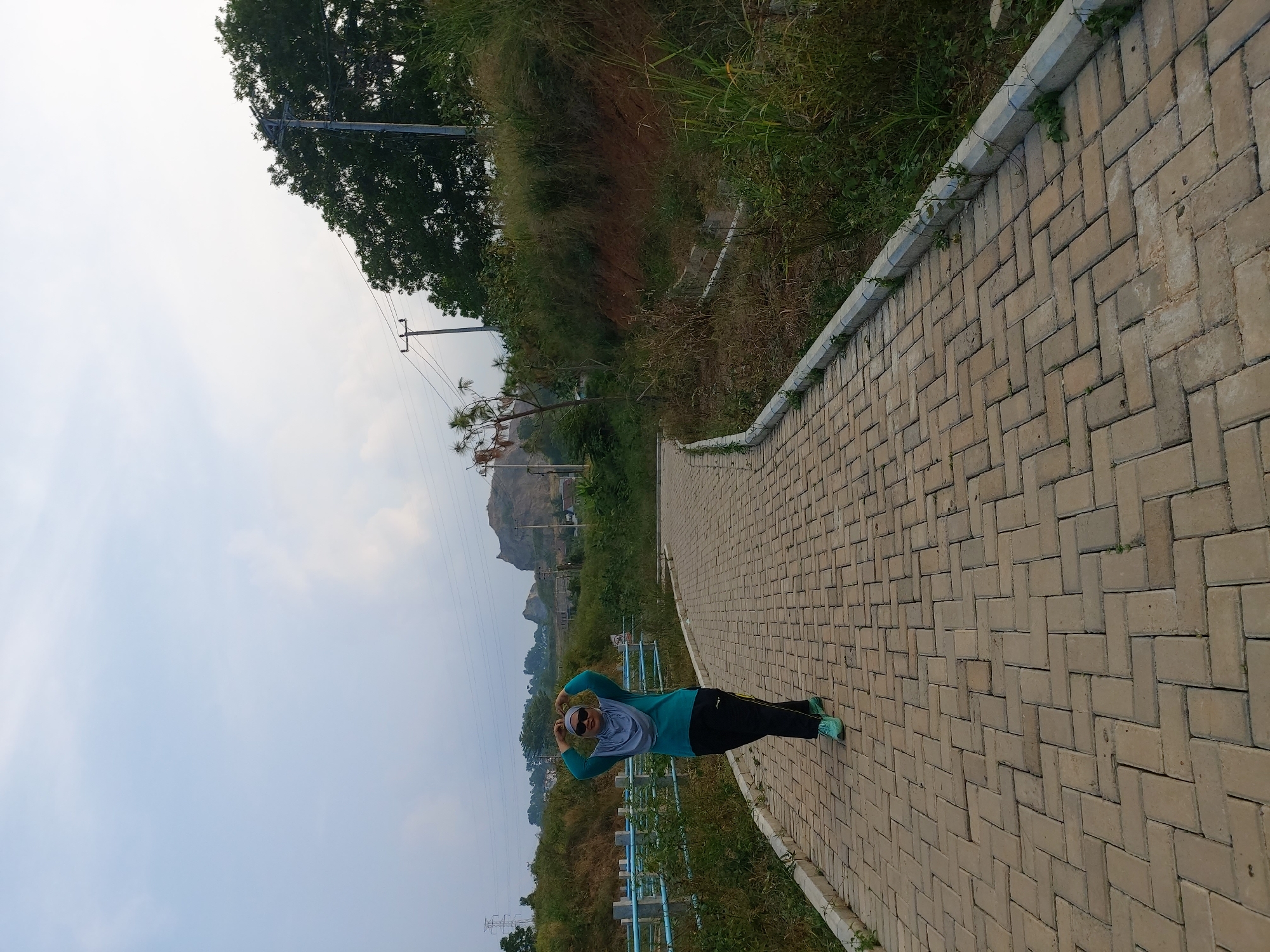 A woman wearing a teal long-sleeved shirt, a gray hijab, black pants, and turquoise sneakers is standing on a paved walkway. Her hands are raised to her head, forming a heart shape. She is wearing sunglasses. The walkway is made of beige rectangular pavers laid in a herringbone pattern. The walkway curves to the right. To the left of the walkway, there is a blue metal railing and lush green vegetation. Beyond that, there is a brown embankment. To the right of the walkway, there is a dirt embankment covered in dry grass and some green plants. In the background, there are trees, utility poles with power lines, and a rocky hill with some buildings. The sky is light blue with some white clouds. The overall scene appears to be an outdoor recreational area. The location is not explicitly identifiable as Bandar Lampung, Indonesia, based solely on the visual information.
