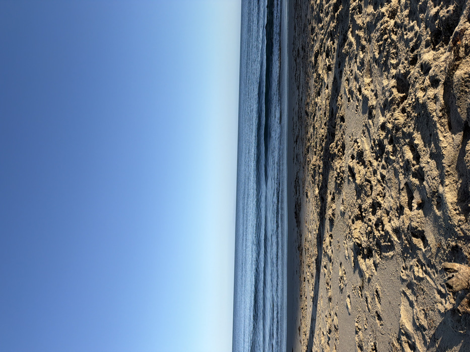 The image depicts a clear blue sky over the Pacific Ocean and a sandy beach in Malibu, United States of America. The sky, occupying the upper portion, is an uninterrupted gradient of light to medium blue. The ocean occupies the mid-ground, displaying multiple parallel wave sets of varying intensity, with darker blue water extending towards the horizon line. The foreground consists of textured, light brown sand with numerous depressions and footprints, indicative of recent activity. A distinct shadow extends across the lower left quadrant of the sand. No people, specific objects, or ongoing actions are visible in the scene.