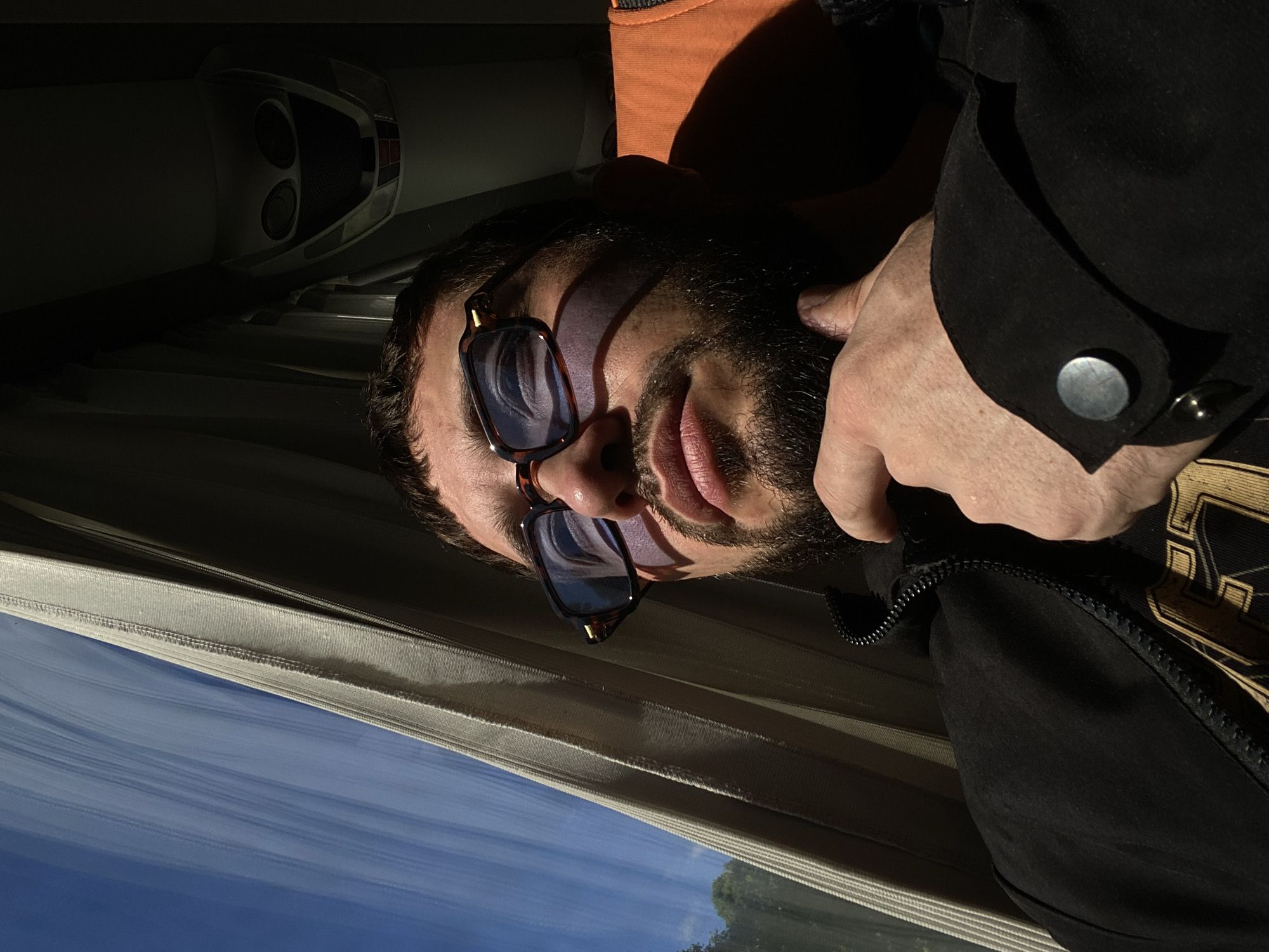 A man is seen from the chest up, wearing sunglasses and a black jacket. He is resting his chin on his right hand. The man is looking to the left, out a window that is covered by a beige curtain. The window shows a bright blue sky with some white clouds and trees. The man's face is illuminated by sunlight, casting a shadow on the right side of his face. The background is dark, with what appears to be part of a bus or train interior visible, including an orange seat. The scene is lit by natural light coming from the window. There is no discernible city or location mentioned in the image or its context.
