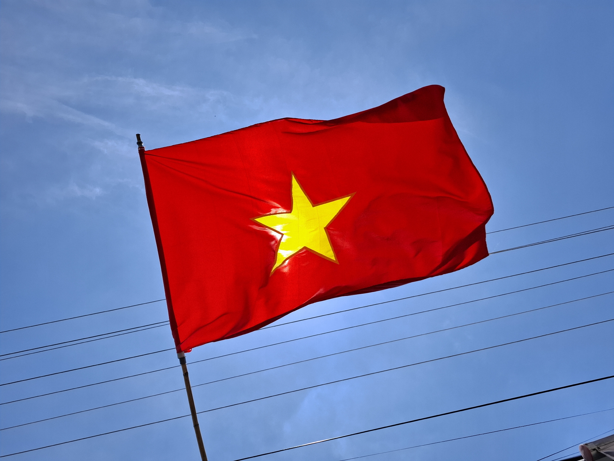 The image shows the national flag of Vietnam waving against a clear blue sky with scattered white clouds. The flag is red with a large yellow five-pointed star in the center. It is attached to a bamboo pole, which is strung with several dark electrical wires that stretch across the frame. The lower left corner of the image shows a small portion of what appears to be a building. The photograph was taken from a low angle, looking up at the flag and sky. The scene suggests an outdoor setting, possibly in a residential or urban area within Phu Thuy Ward, Vietnam, given the presence of electrical wires and a building. There are no people or animals visible in the image. The lighting indicates it is daytime.