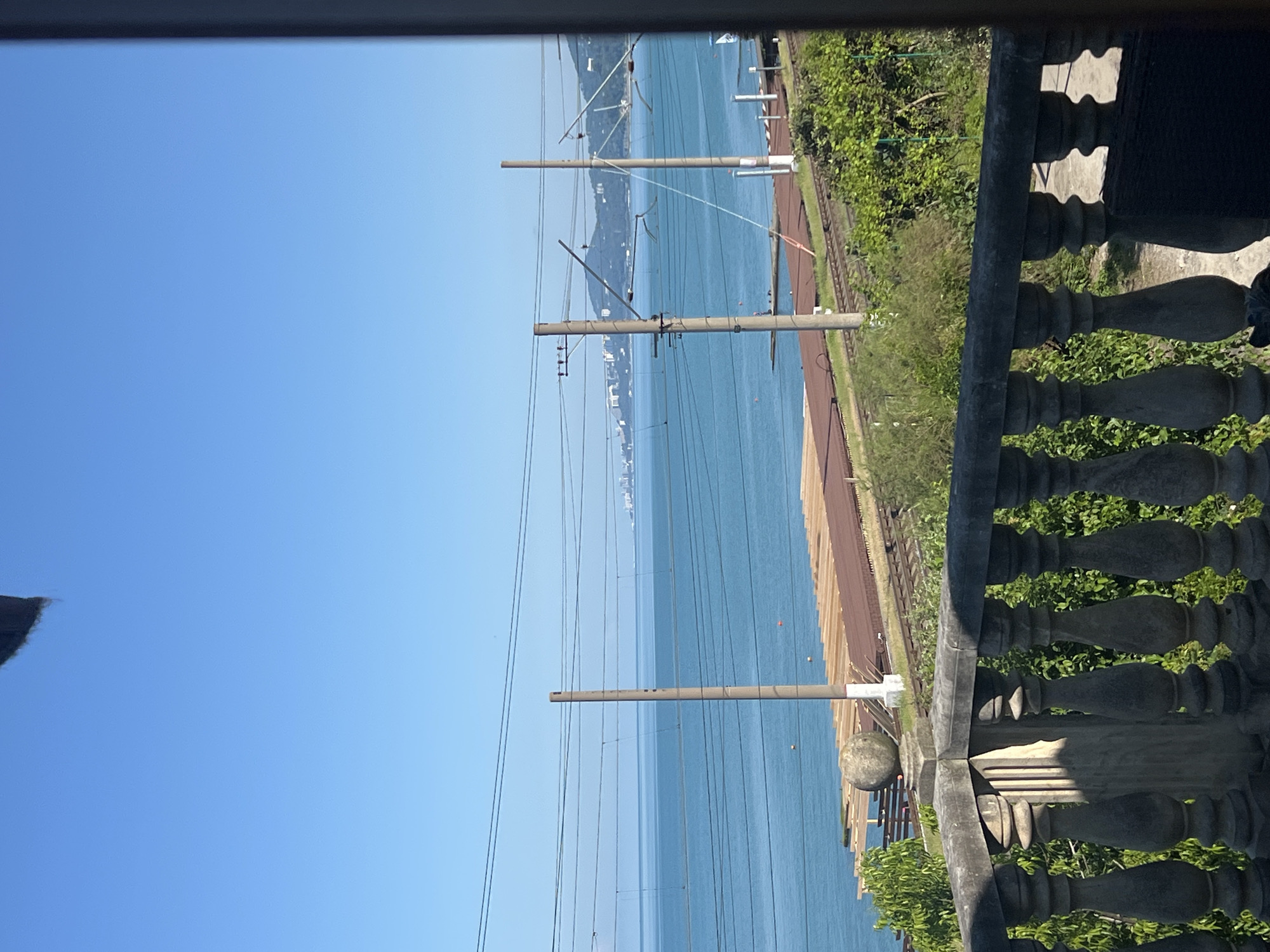 The image shows a view of the sea from a balcony. In the foreground, a stone balustrade is visible, with decorative pillars and a carved top railing. Behind the balustrade, lush green foliage can be seen. Further back, there is a railway line with overhead power lines and support poles. Beyond the railway tracks, the blue sea stretches out to the horizon, where a distant city skyline and hills are visible under a clear blue sky. The overall scene depicts a coastal urban environment on a sunny day.