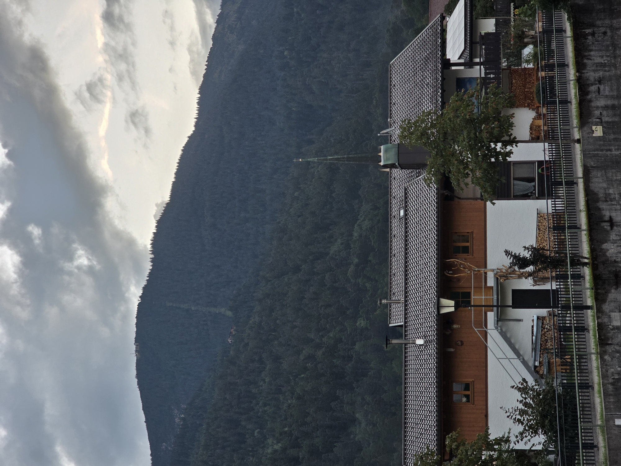 The image depicts a view of buildings nestled at the base of a densely forested mountain, under a cloudy sky. The buildings appear to be residential, with dark tiled roofs, white and brown painted facades, and visible windows and doors. A church steeple with a cross is visible rising above the rooftops. In the foreground, a dark metal fence runs horizontally across the image, separating the buildings from a concrete embankment. A lamppost stands near a set of outdoor stairs leading to one of the buildings. Firewood is stacked in piles outside some of the structures. The mountain behind the buildings is covered in a dark green forest, with a visible track or path ascending its slope. At the very top of the mountain, a cluster of structures and what appears to be a ski lift or tower are visible. The overall atmosphere is overcast and slightly muted in color. This scene is consistent with the scenery found in Garmisch-Partenkirchen, Germany, a town known for its mountainous terrain and winter sports facilities.