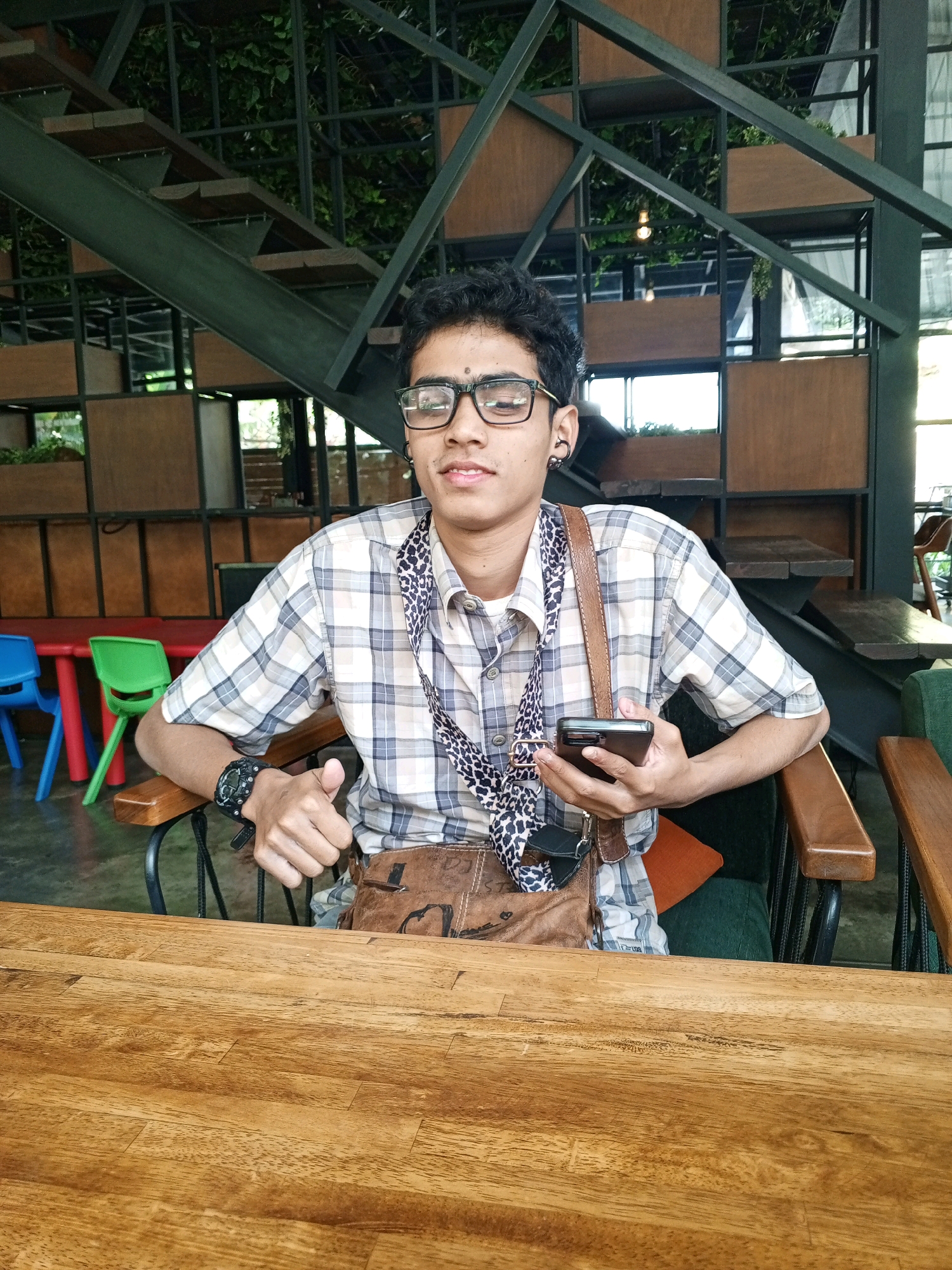 The image depicts a young man sitting at a wooden table in what appears to be a cafe. He is wearing a plaid shirt, glasses, and a crossbody bag. He is holding a smartphone in his right hand and giving a thumbs-up with his left hand. Behind him, a modern staircase with wooden steps and metal railings is visible, along with shelves and greenery, suggesting an indoor-outdoor space. The presence of colorful chairs and tables in the background indicates a public seating area. The setting is likely a cafe in Denpasar, Indonesia, characterized by its contemporary design and ample natural light.