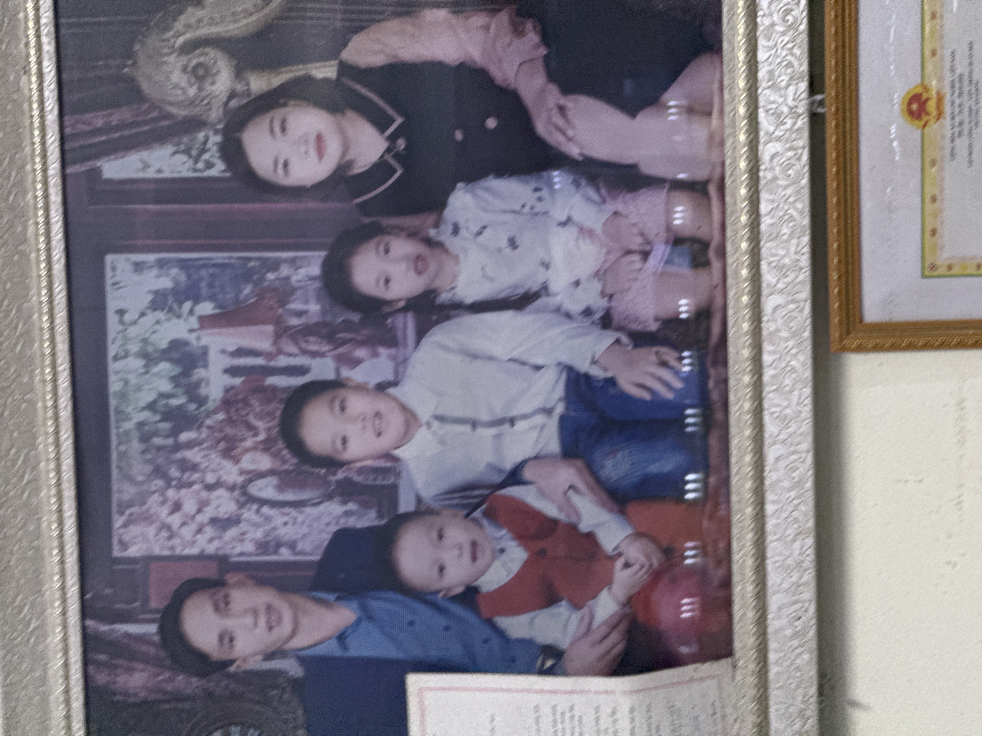 A framed family portrait hangs on a wall, likely inside a home, possibly in Du Già Commune, Vietnam. The portrait depicts a family of five: a mother, father, and three children. The mother is at the top, dressed in a dark top with a light-colored collar. The father is at the bottom, wearing a blue shirt. The three children are positioned in between. The youngest child, dressed in a red vest, is in the father's arms. Behind the family is a painted backdrop with a decorative chair and a landscape. Adjacent to the family portrait is another framed document with an ornate border. The wall is a plain off-white color.