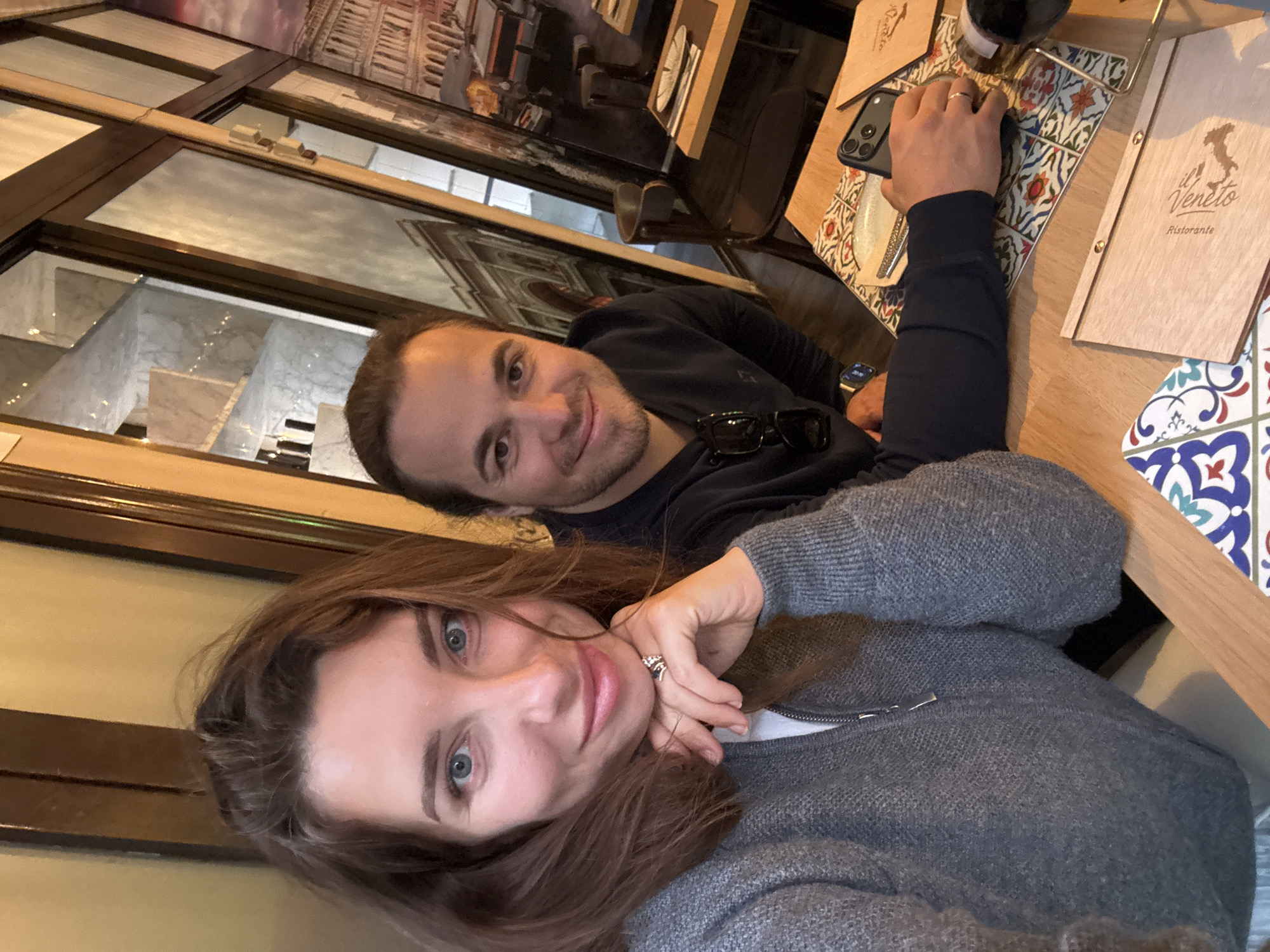 The image captures two individuals, a woman and a man, seated at a wooden table inside a restaurant in València, Spain.
In the foreground, a woman with long brown hair and blue eyes looks directly at the camera with a soft smile. She is wearing a gray knitted cardigan with a zipper and has a ring on the fourth finger of her left hand. Behind her, a man with dark hair and a beard also smiles towards the camera. He is wearing a dark long-sleeved top, with sunglasses resting on his chest and a watch on his left wrist. His right hand is placed on a smartphone (an iPhone) that rests on a colorful, patterned tile placemat on the table.
The scene suggests an activity of dining or socializing in a restaurant. On the wooden table, alongside the placemat and phone, there is a wooden menu booklet prominently featuring the text "il Veneto Ristorante" with an outline of Italy. The placemat displays a vibrant mosaic-like pattern in hues of blue, red, orange, and green. The background reveals reflections in large glass partitions, showing an interior space with stairs and marble finishes. Beyond the reflections, wall decor depicts architectural murals, possibly evoking Italian cityscapes. The lighting suggests it is either daytime or early evening, creating a relaxed and pleasant atmosphere, with both subjects appearing content.