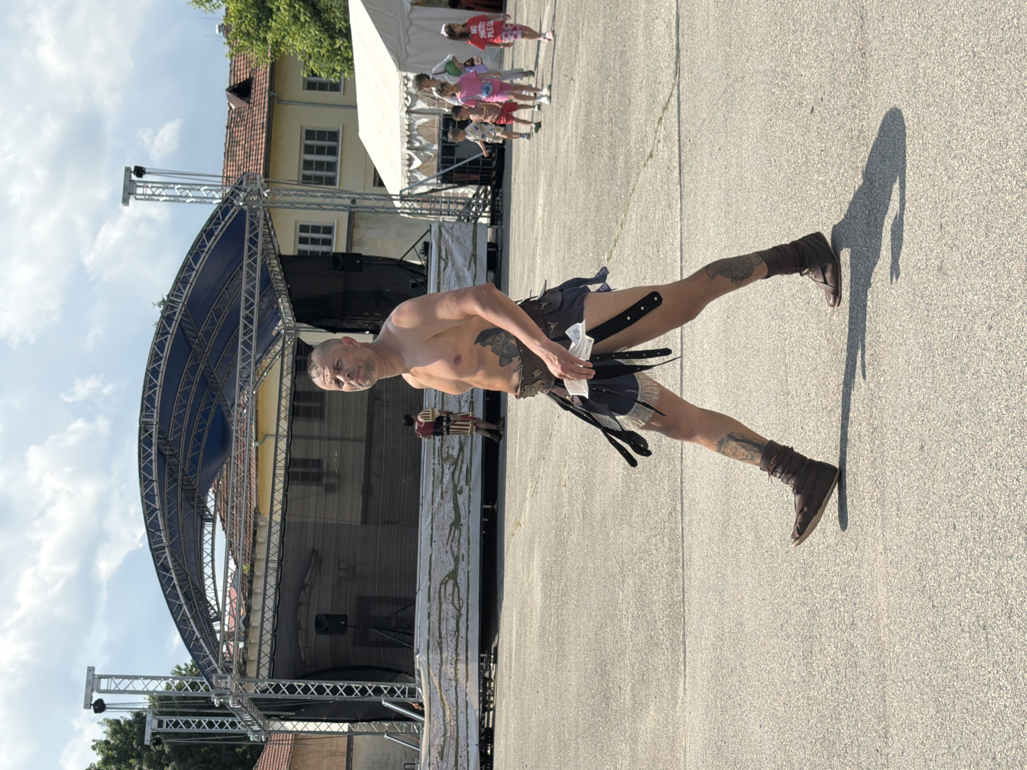 The image captures an outdoor scene in Pleven, Bulgaria, under a partly cloudy sky with bright sunlight, indicated by pronounced shadows. In the foreground, a shirtless adult male with a shaved head, visible chest and arm tattoos, walks rightward across a light gray paved surface. He wears a dark brown leather belt with metallic details, a dark blue kilt-like garment with fringe and black straps, and dark brown laced ankle boots. He holds white papers in his left hand.
In the mid-ground, a large outdoor stage structure with a blue arched canopy and silver metal trusses dominates the left side. Speakers are mounted on the trusses, and a wide dark banner, featuring a light-colored, map-like design, stretches across the stage front. Behind this banner, another individual in a historical costume (dark red vest, striped lower garment) is visible. To the right of the stage, a light-colored building with multiple windows and a red tiled roof stands. Further right, a white tent is visible. A group of four children and one adult female walk towards the viewer on the paved surface, positioned near the white tent. One child wears a red t-shirt with "NO PHOTO PLEASE" printed on it.