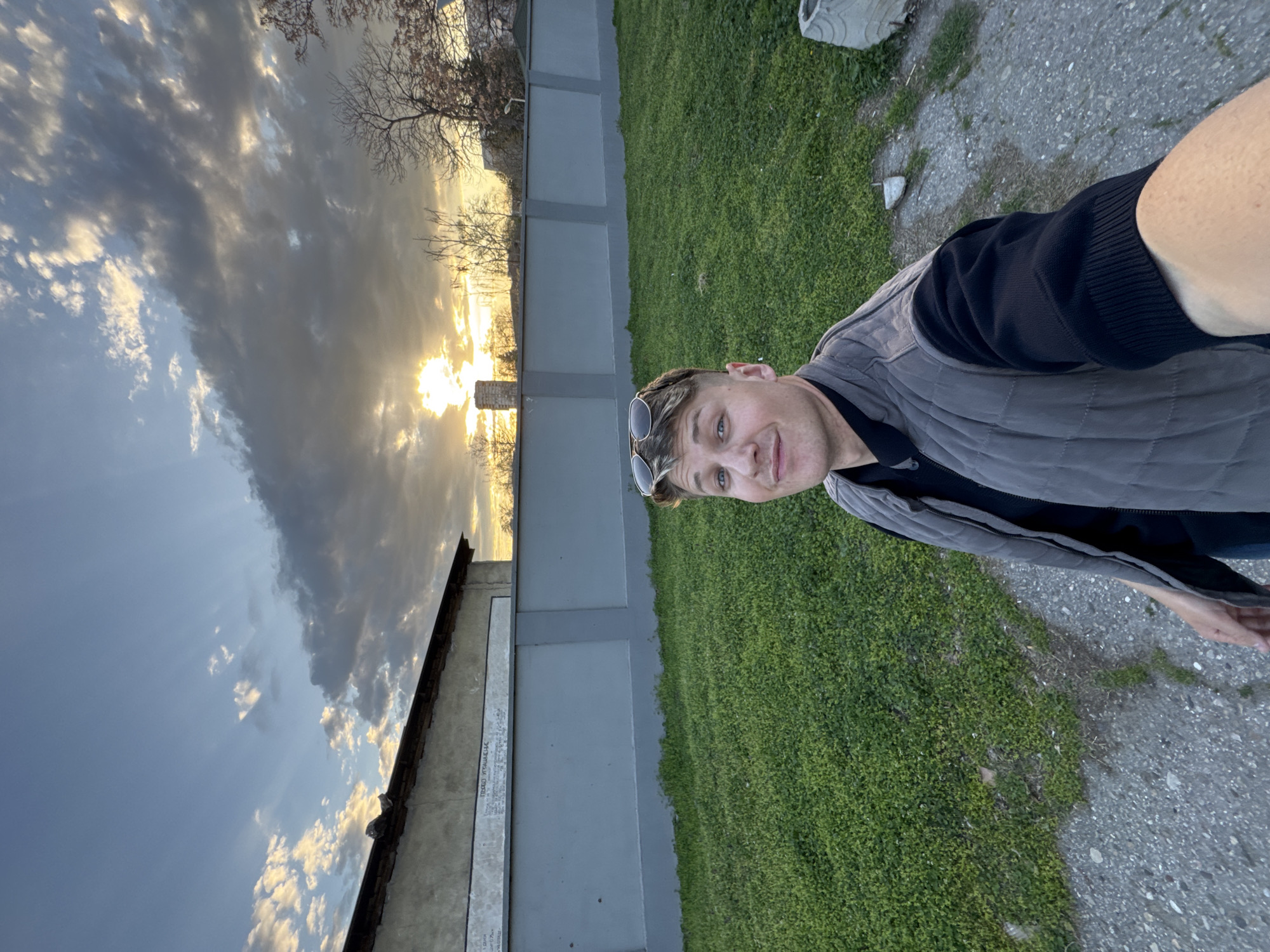 The image is a selfie of a man standing next to a gray fence, with grass on the right and a structure with writing on the left. The sky is visible in the background, with clouds and a sunset. The picture was taken in Plovdiv, Bulgaria. The structure with writing is the Brezovska Street Massacre Memorial located in Plovdiv. The man is wearing a gray vest and sunglasses on his head. He's on a sidewalk.