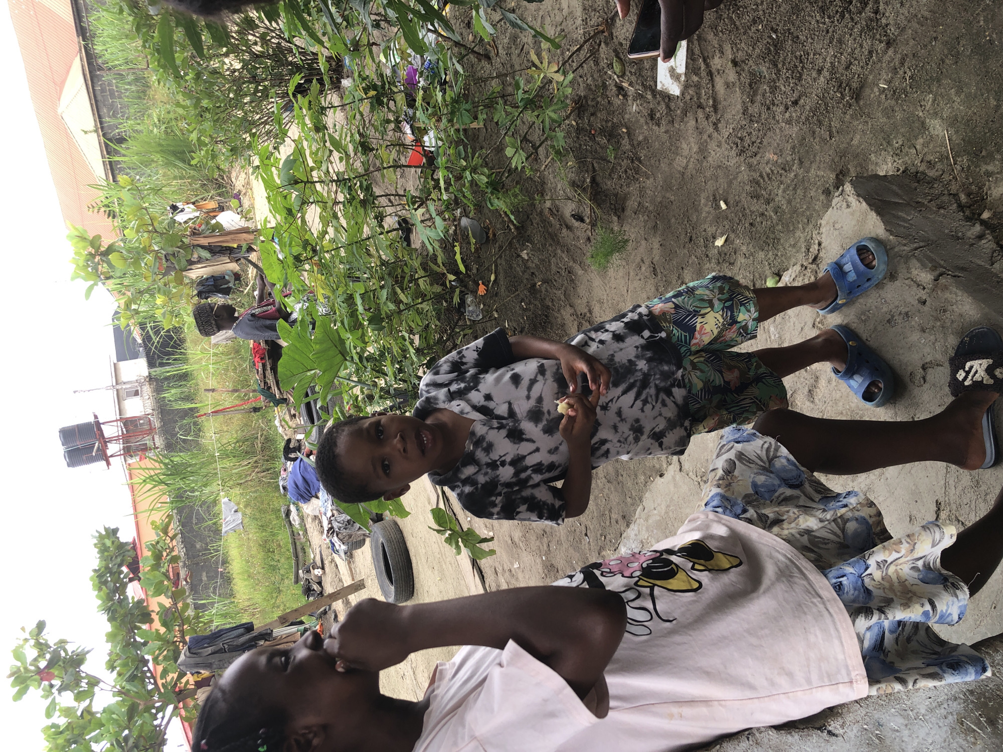 The image depicts an outdoor scene in Ado, Nigeria. In the foreground, two children are visible. A girl on the left, wearing a light pink t-shirt with a cartoon character and a floral skirt, is turned to the side. A boy on the right, in a black and white tie-dye t-shirt and colorful shorts, is facing forward and holding a piece of food. The ground is dirt and sand, with some green plants and a concrete step. In the background, there is a fence, buildings, and a water tank on a stand. Various objects, including clothes and tires, are scattered around the area, suggesting a lived-in environment. The overall impression is a candid moment captured in an informal setting.