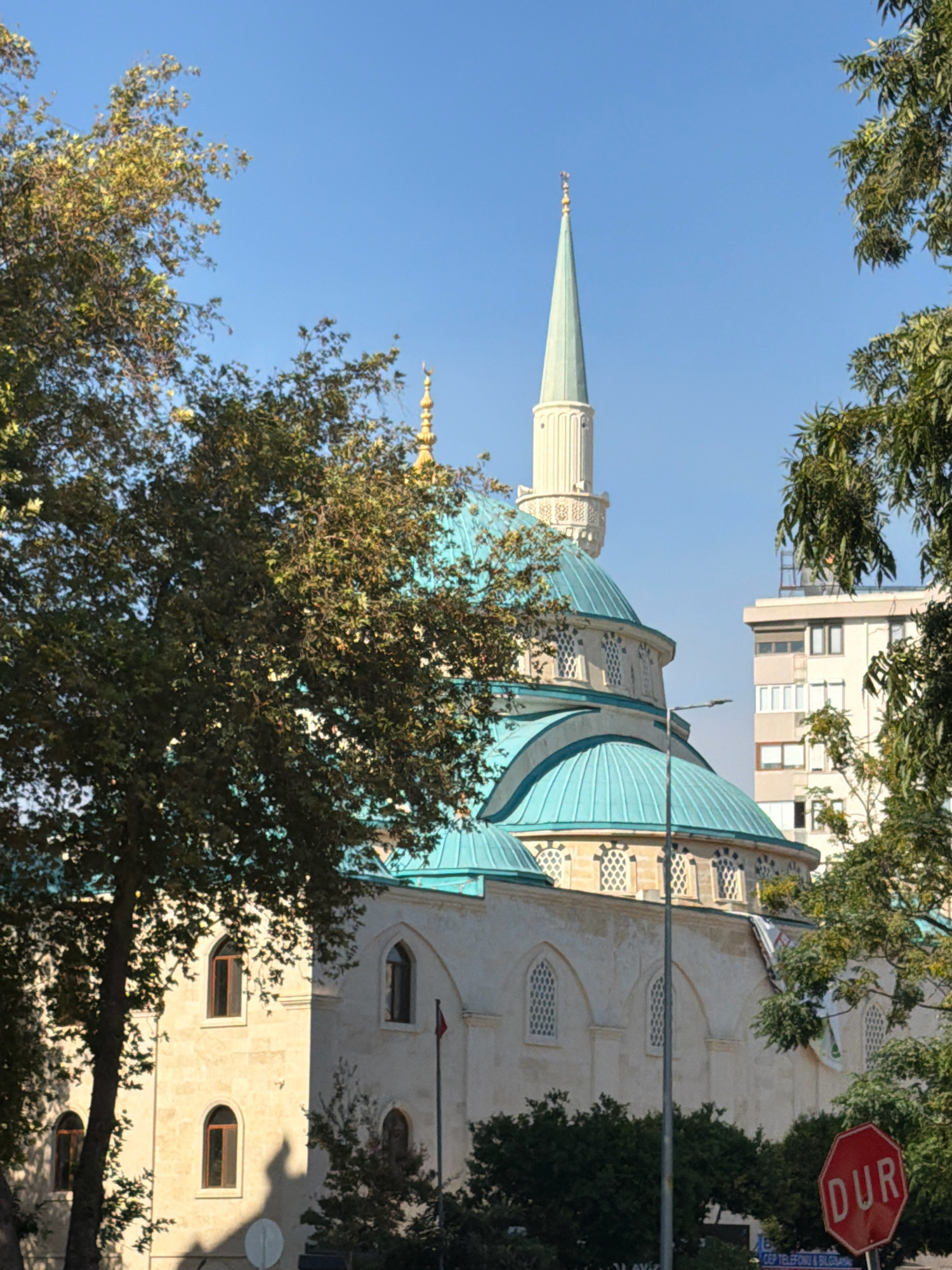 The image captures a section of the Yivli Minareli Mosque in Antalya, Turkey, seen through the foliage of trees on a clear, sunny day. The mosque features a light-colored stone facade with arched windows and multiple turquoise domes. A tall, slender minaret, also light-colored with intricate detailing, rises prominently from the structure. In the background, a modern multi-story building stands partially visible. In the foreground, a hexagonal red stop sign with the word "DUR" (Turkish for "stop") is positioned at the bottom right of the frame. The overall scene depicts an urban landscape where historic architecture is integrated with modern city elements and natural vegetation.