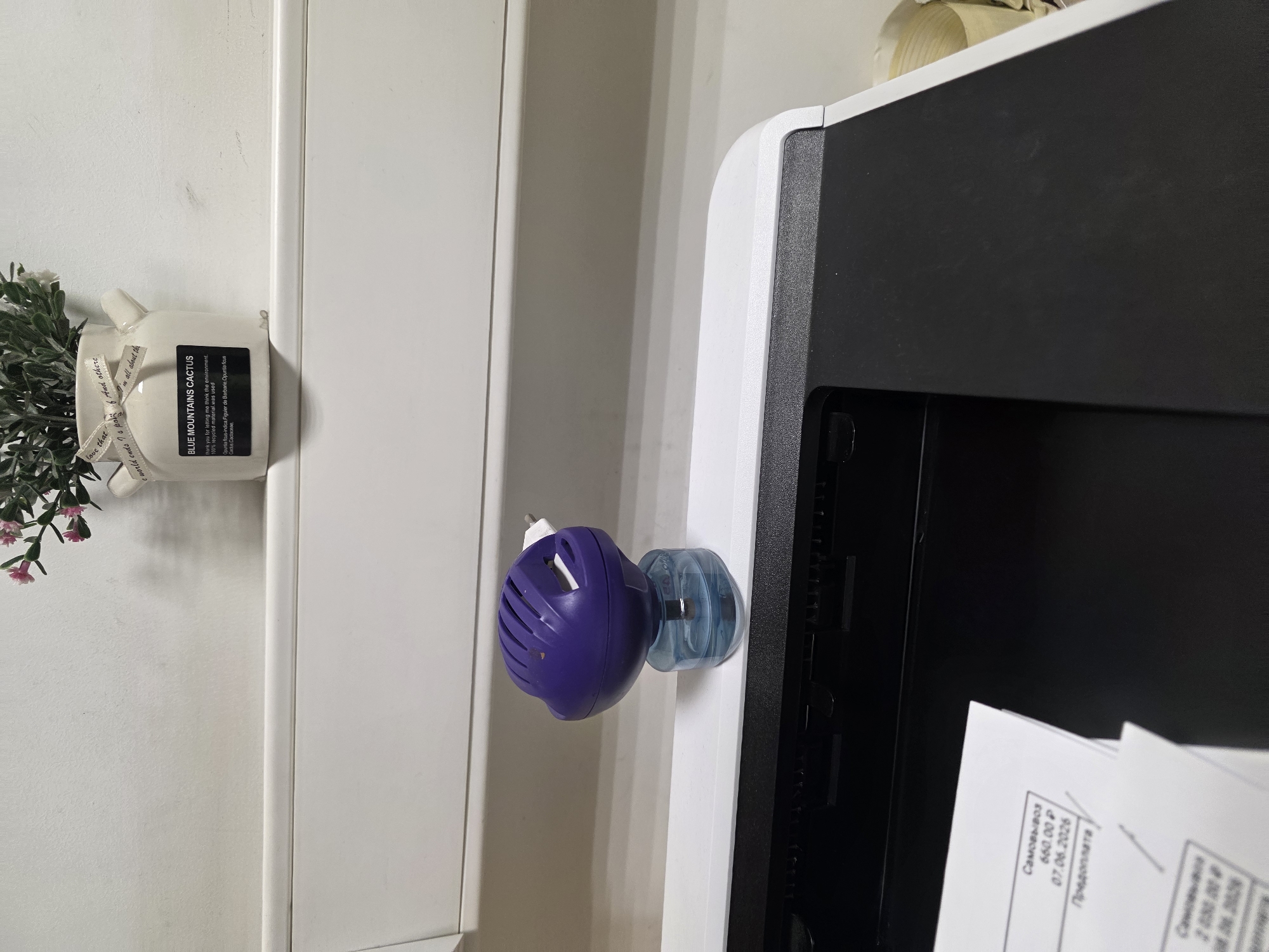 A close-up shot focuses on a purple electric air freshener plugged into a white shelf, positioned above a black and white printer. To the left, a corner of printed documents is visible. In the upper background, a small white vase with green artificial plants and pink flowers sits on the shelf, adorned with a ribbon that has text printed on it. The overall scene is indoors, with the background appearing to be a plain white wall.