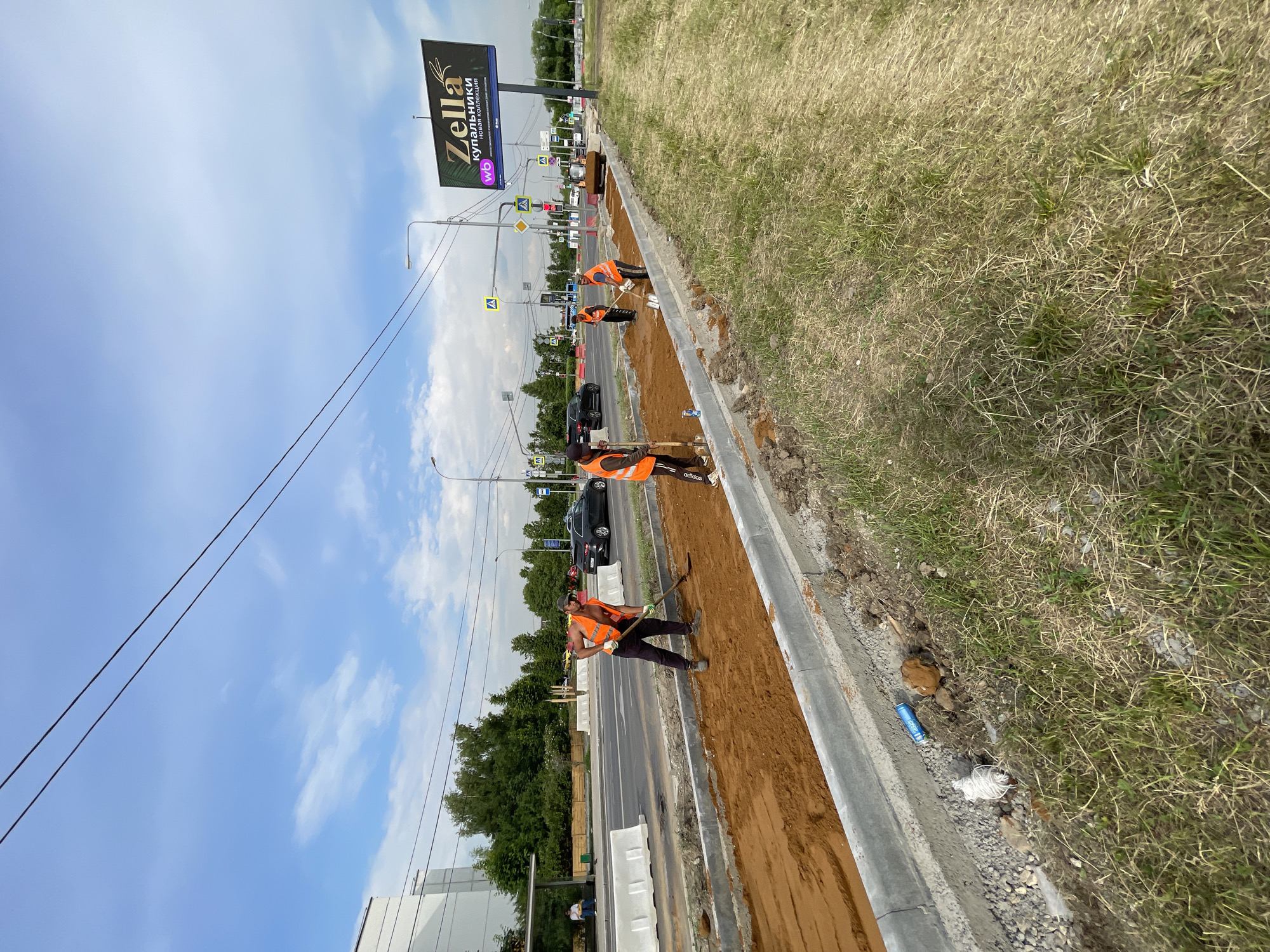 The media file depicts an outdoor scene on a bright, overcast day in Moscow, Russia, likely during daytime. The primary subjects are construction workers engaged in road or sidewalk repair.
Several individuals are visible, all wearing bright orange safety vests, indicating their work attire. They are using shovels to move and level a reddish-brown soil or sand material on what appears to be a construction site adjacent to a road. One worker, with his vest unzipped, is actively shoveling. Another, further down the line, is also working with a shovel. Two more figures in vests are further up the construction area.
The setting is a roadside with a paved road to the left, featuring several cars, including a black sedan. Traffic lights and road signs are visible in the background. To the right of the construction area is a grassy verge, with dry, straw-like grass, suggesting a warm season. Beyond the road, there is a line of trees. A large billboard with the text "Zella купальники новая коллекция" and a stylized logo featuring the letters "wb" is prominently displayed in the background. The sky is a light blue with scattered white clouds.
Notable details include the construction activity, the presence of vehicles on the road, and the clear signage of a commercial advertisement. The overall impression is one of ongoing urban infrastructure work under typical daylight conditions.
