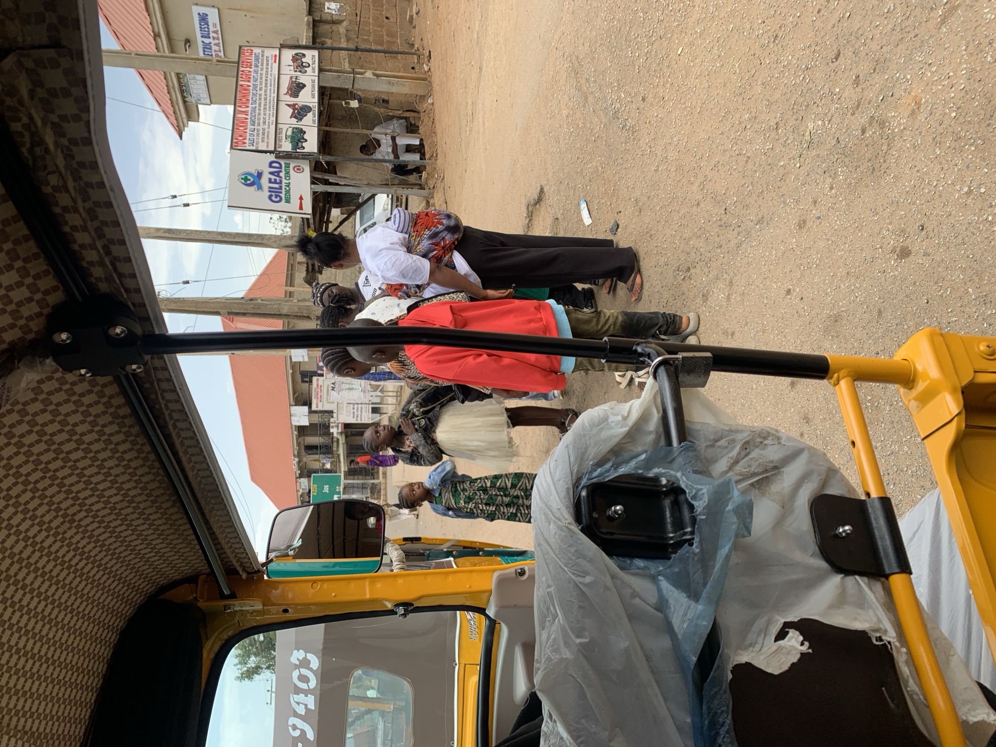 The image, taken from inside a yellow motorized tricycle in Rafin Par, Nigeria, captures a street scene. Several individuals, mostly children, are standing or walking alongside the road. Some appear to be looking towards something out of the frame, perhaps interacting with someone or something not visible. The adults are dressed in ordinary clothes, while the children wear dresses or casual shirts.
Behind the people, street signs and buildings are visible. One sign reads "TOCHUKWU IK OKONKWO AGRO SERVICES SALES OF ALL AGRICULTURAL TRACTORS SPARE PARTS AND IMPLEMENTS, RAFINPAR HEAD OFFICE MARIN GADA. JET IVECO TRUCK." Another sign indicates "GILEAD MEDICAL CENTRE". The buildings have simple architectural features. The street appears to be unpaved and sandy.