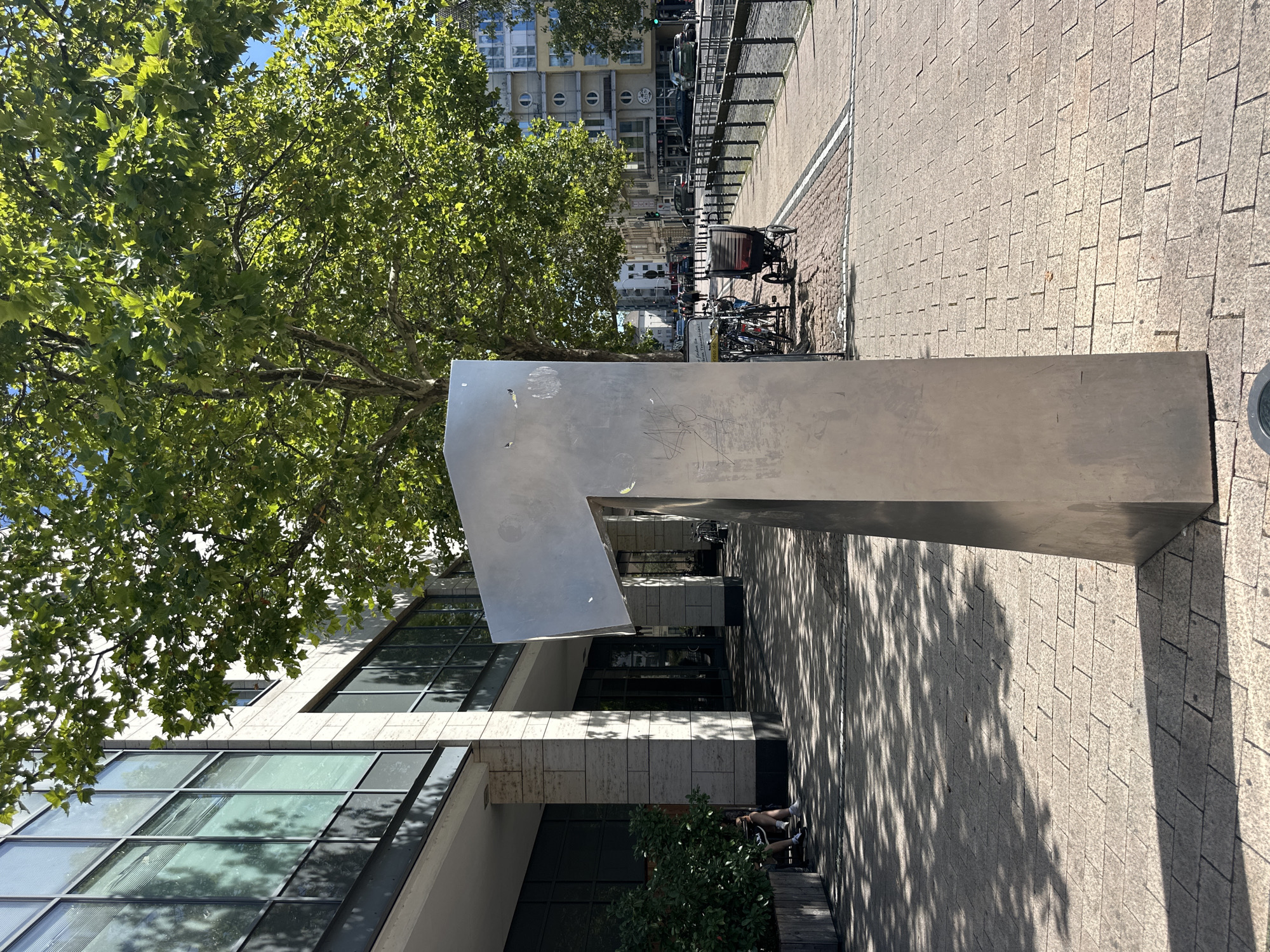 The image depicts a street scene in Cologne, Germany, with a large, metallic sculpture resembling the number "1" in the foreground. The sculpture is positioned on a paved sidewalk. To the left of the sculpture is the facade of a modern building with large windows and stone cladding. Some of the windows have blinds. A person is seated on a low wall or planter box near the building entrance, partially obscured by foliage.
The sidewalk is made of interlocking bricks and is partially covered in dappled sunlight and shadow from trees. Further down the sidewalk, several bicycles are parked. A stroller or small cart is also visible.
The background reveals a street with traffic, including cars and a bus. Buildings with diverse architectural styles line the street, some featuring circular windows. A black metal railing runs along the edge of the sidewalk, separating it from the street. The sky is blue and clear. The overall impression is that of a sunny day in an urban environment.