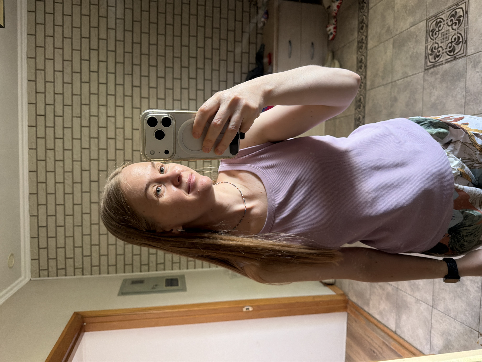 A woman is taking a selfie in a bathroom. She has long blonde hair and is wearing a light purple tank top and patterned shorts. She is holding a smartphone in her right hand and looking at its reflection in a mirror. The background shows a brick wall, a doorway, a chest of drawers, and a tiled floor. The woman is in Khabarovsk, Russia.