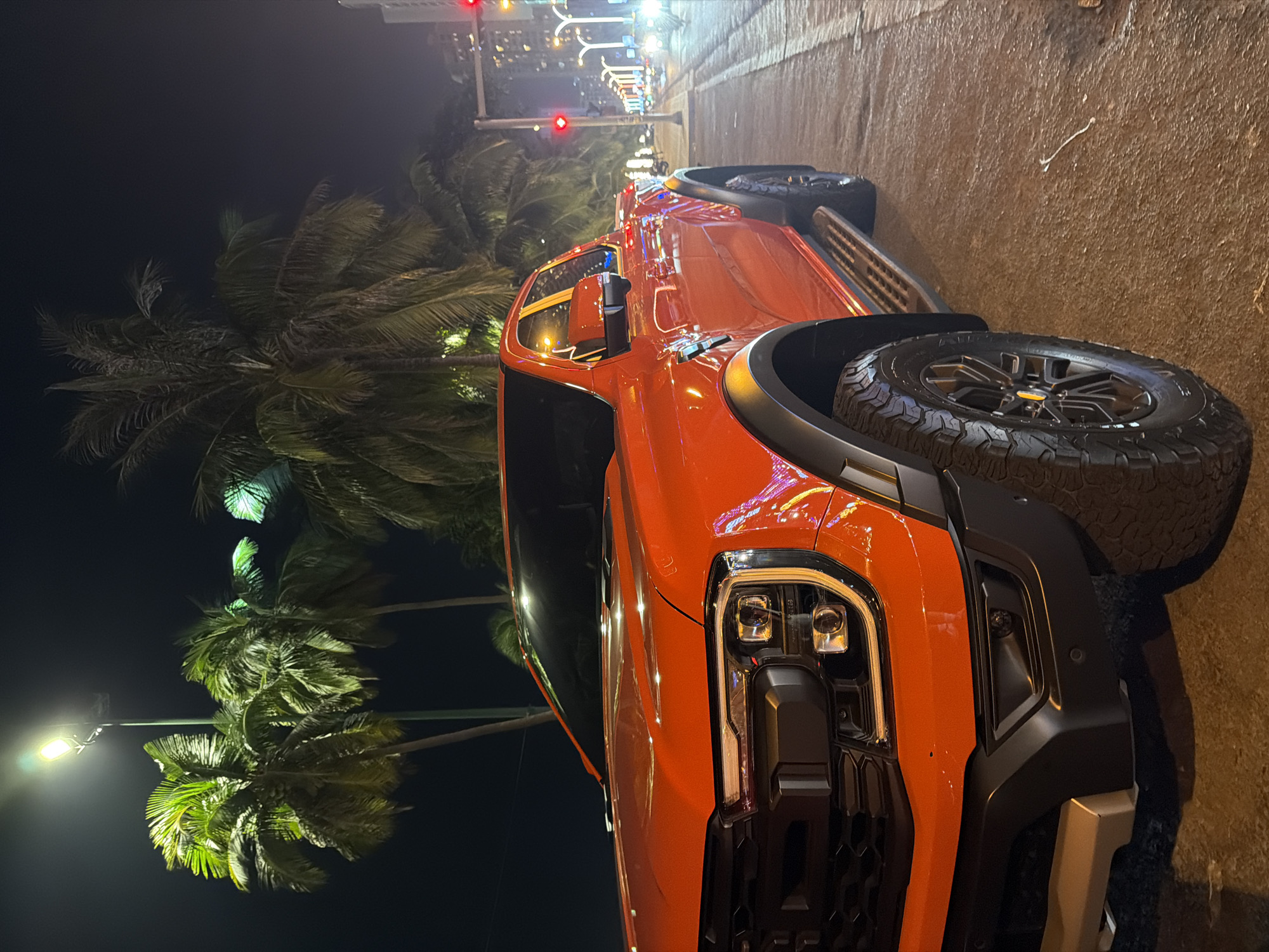 The image shows a bright orange Ford Ranger Raptor pickup truck parked on a city street at night. The truck is positioned at an angle, with its front left side prominently featured. The background reveals a street scene with palm trees, streetlights, and buildings illuminated by artificial lights. Traffic lights are visible in the distance, one of which is red. The street appears to be wet, possibly from rain, and reflects the surrounding lights. The overall setting suggests an urban environment, likely in a tropical or subtropical location due to the presence of palm trees. The image was taken in Nha Trang, Vietnam.