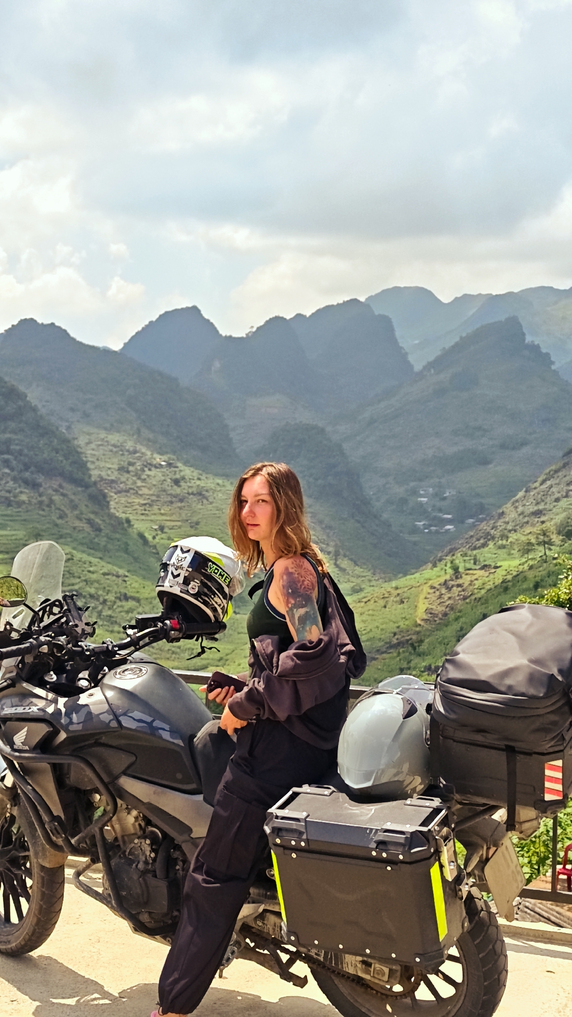 A young woman is sitting on a motorcycle in Lũng Phìn Commune, Vietnam. She is turned toward the camera and has brown hair and a tattoo on her left arm. She is wearing dark pants and a dark tank top, with a dark jacket draped around her shoulders. She is holding a phone in her hands. The motorcycle is black and has luggage attached to the rear. A helmet is sitting on the front of the motorcycle. The background consists of green, mountainous terrain under a partly cloudy sky.