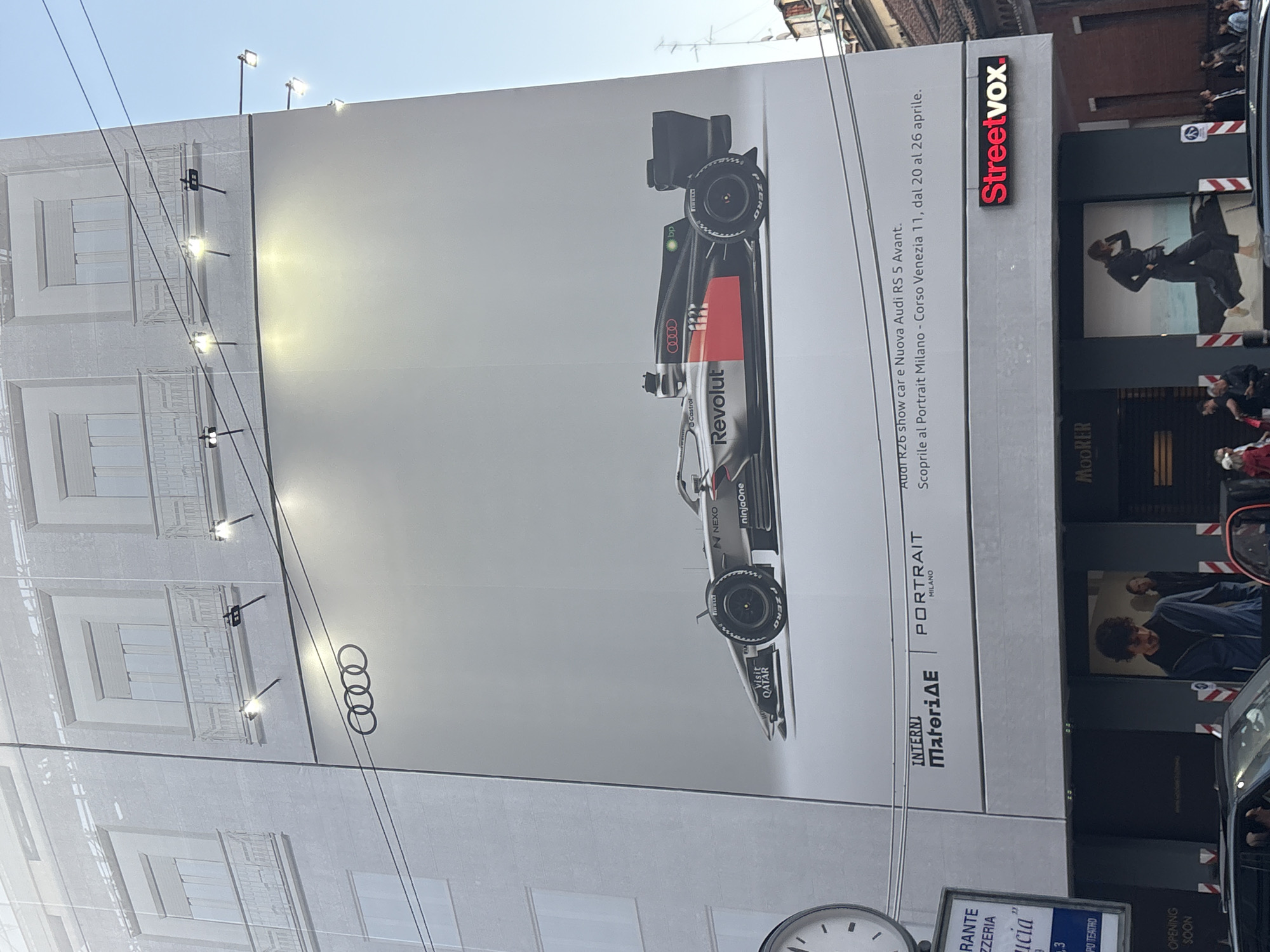 A large, vertically oriented billboard dominates the left side of the frame, attached to a building facade in an urban setting. The billboard features a sleek, black, white, and red Audi R26 F1 show car, adorned with various sponsor logos including "Revolut," "bp," "Castrol," "NEXO," "ninjaOne," and "Visit QATAR," as well as "P ZERO" on the tires. The iconic Audi rings logo is also visible on the billboard.
The text on the billboard advertises an event: "Audi R26 show car e Nuova Audi RS 5 Avant. Scoprile al Portrait Milano - Corso Venezia 11, dal 20 al 26 aprile." (Discover the Audi R26 show car and new Audi RS 5 Avant at Portrait Milano - Corso Venezia 11, from April 20 to April 26.) Additional text includes "INTERNI MateriaAE" and "PORTRAIT MILANO."
To the right of the billboard, a section of another building or storefront is visible, featuring signs for "Streetvox" and "MOORER." Window displays with mannequins are present in these storefronts. The street level shows several pedestrians, partially obscured by what appears to be a parked car on the far right.
The overall scene suggests a busy daytime environment, with clear skies. Several spotlights are mounted on the top and side of the billboard structure, and power lines or cables stretch across the upper left portion of the image. A street clock is partially visible at the bottom left, displaying "OPENING SOON" and fragments of Italian words like "RANTE," "ZZERIA," "icia," and "PO TEATRO," likely from a separate sign below. The architecture and text confirm the location as Milan, Italy.