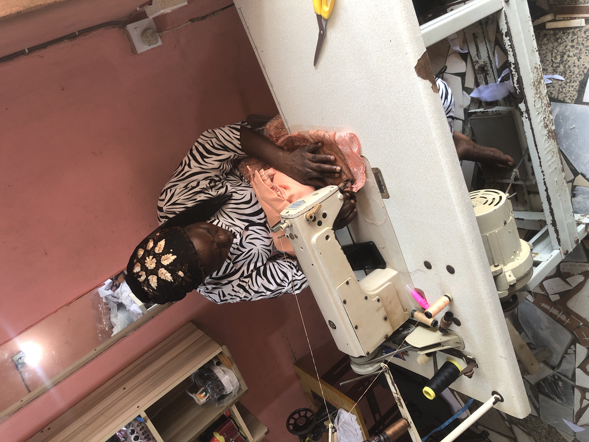 A woman with a black and white zebra-print shirt and a black headscarf adorned with gold leaf-like embellishments is operating a beige sewing machine. She is focused on the fabric in front of her, which is a peachy-orange color with intricate embroidery. The sewing machine is placed on a large, white table. To the right of the sewing machine, a pair of yellow-handled scissors rests on the table. The background of the scene features a pink wall. To the left, there is a wooden shelf unit with various items on it, including spools of thread and what appears to be packaged goods. The floor is tiled with a mosaic pattern of brown, gray, and white tiles. The overall setting suggests a sewing workshop or tailoring shop. The presence of electrical outlets and the general state of the room indicate an urban environment, possibly within a residential or commercial space in Ado, Nigeria.