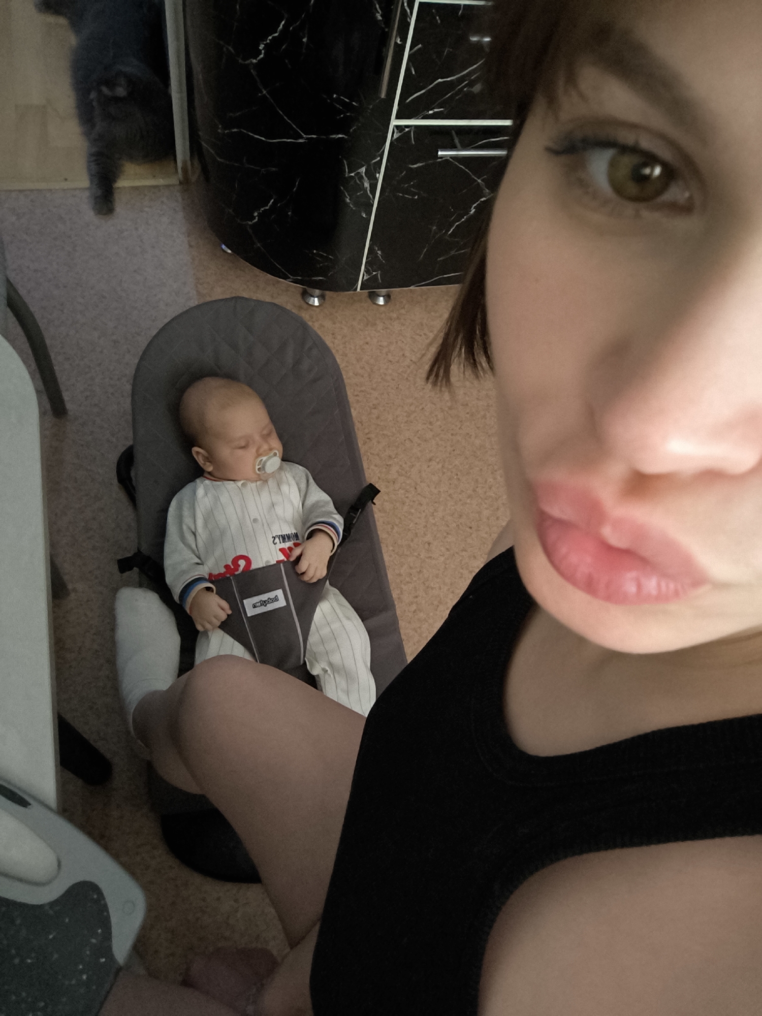The image shows a close-up selfie of a woman with her baby and a cat in the background, set indoors in Chaikovsky, Russia. The woman's face is prominent on the right side of the frame, showing her right eye, nose, and lips pursed in a slight pout. She has brown hair and light brown or hazel eyes, and wears a black top.
To the left and slightly below the woman, her baby is sleeping peacefully in a gray bouncer, secured by a harness. The baby has a pacifier in its mouth and is dressed in a light-colored, vertically striped romper. Visible on the baby's chest is reversed text that appears to be "2 YMMOM". The brand name "BABYBJÖRN" is also discernable in reverse on the bouncer's harness. One of the woman's legs, wearing a light-colored sock, is visible near the bouncer.
In the upper left corner of the background, a dark gray cat is partially visible, showing its hindquarters and one leg, seemingly moving away from the viewer. Behind the cat and extending across the upper background is a dark cabinet or piece of furniture with a distinctive white marble-like pattern, featuring silver handles or decorative feet. The floor is a light brown or beige color, appearing to be carpeted or tiled. The scene captures a quiet, domestic moment.