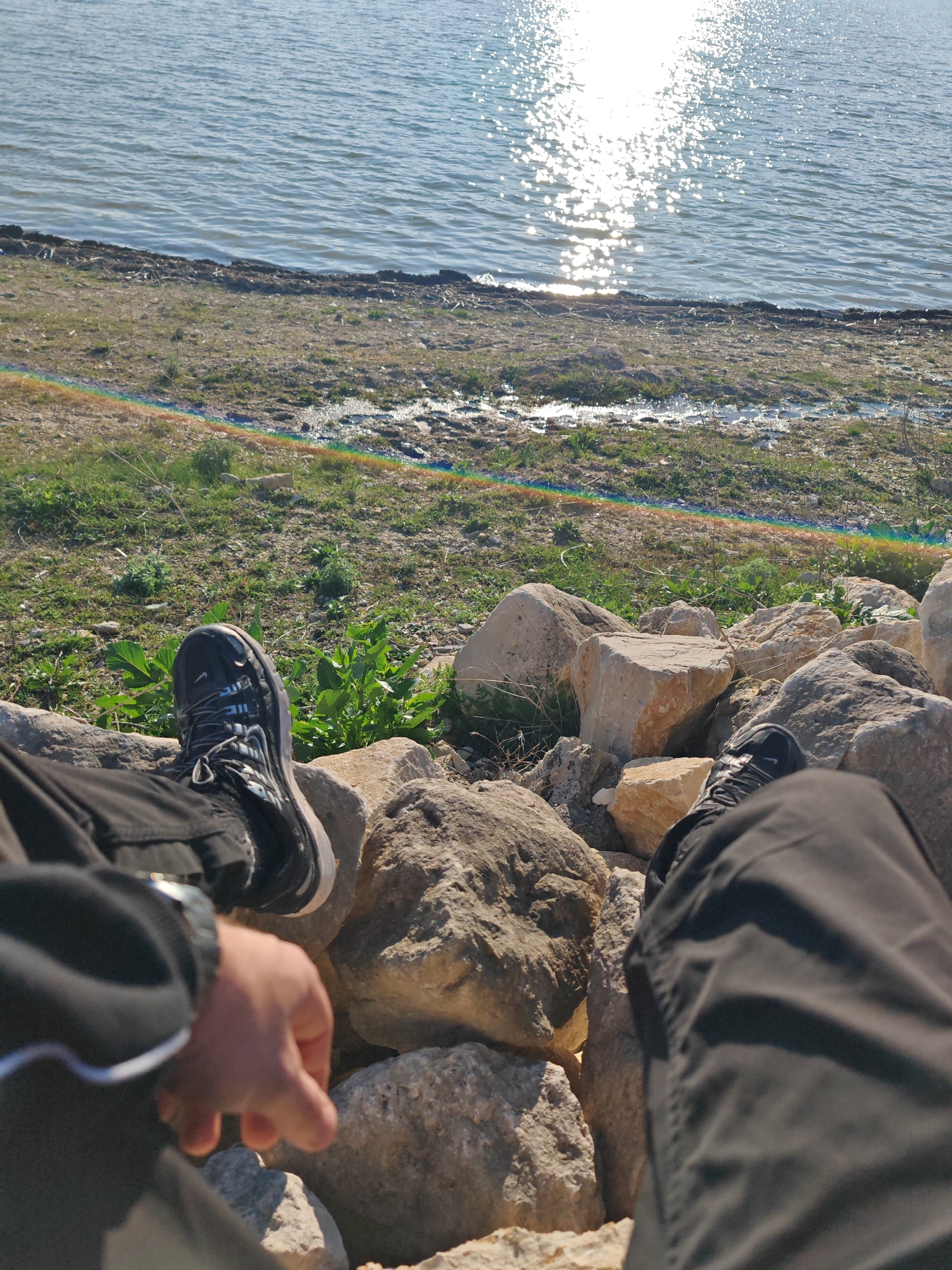A first-person view image shows a person sitting on a rocky bank by a lake, possibly in Shumen, Bulgaria. The person is wearing black shoes and black pants. The lake is in the background with the sun reflecting off the water. The shoreline has grass and some small plants. The rocks the person is sitting on are various sizes and shades of gray and brown. The person's hand and wrist, adorned with a watch, are visible in the foreground.