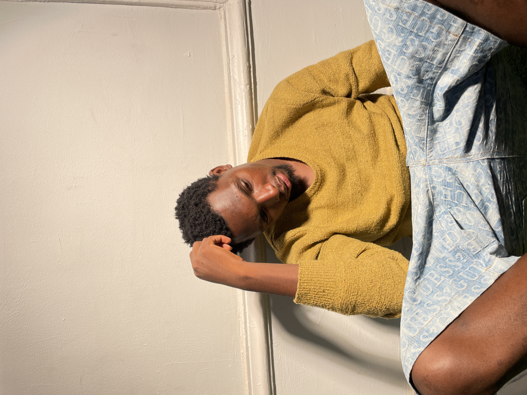 A medium shot captures a dark-skinned man posing indoors in Barakin Akawo, Nigeria. He has short, curly dark hair and a trimmed beard. He is wearing a mustard yellow long-sleeved sweater and light blue shorts patterned with repeated text, which appears to be "GUESS" arranged diagonally. The man is positioned against a textured white wall, with a white door frame visible to his left. His head is slightly tilted, and he looks directly at the camera with a calm, composed expression, his right hand gently resting near his temple. Strong, direct lighting illuminates the scene, casting distinct shadows on the wall and the man, highlighting his features with a warm glow, suggesting bright daylight or a powerful artificial light source. The overall atmosphere conveyed is relaxed and thoughtful.