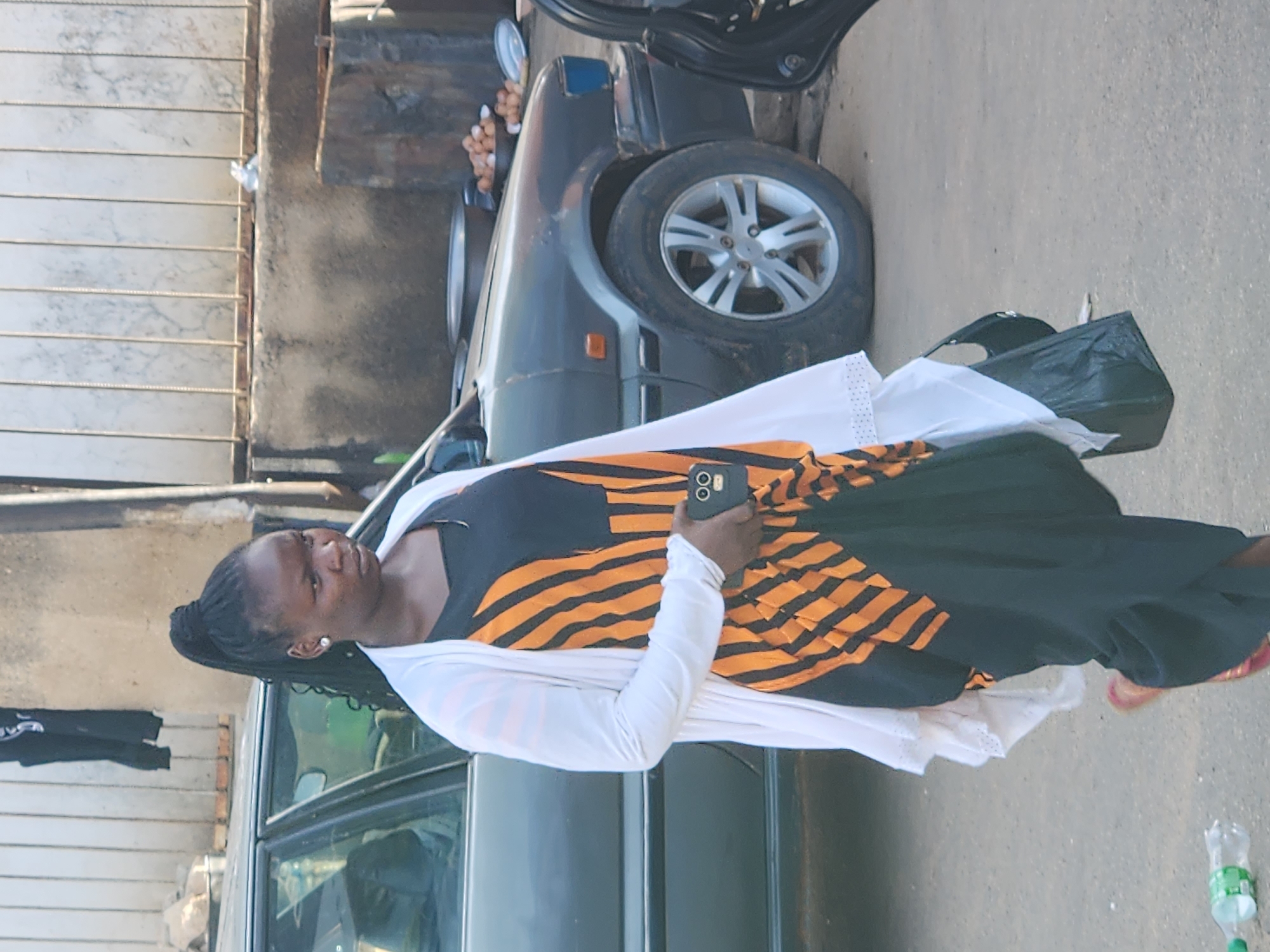 A Black woman walks in front of a parked car. The woman has dark skin and her hair is styled in a high ponytail. She is wearing a white cardigan over a black and orange striped dress, and she is carrying a black bag in her right hand. The car is dark gray and has a blue headlight. In the background, there is a concrete wall with a metal fence on top. A pile of eggs is visible on a ledge near the wall. A plastic water bottle lies on the ground in the foreground.