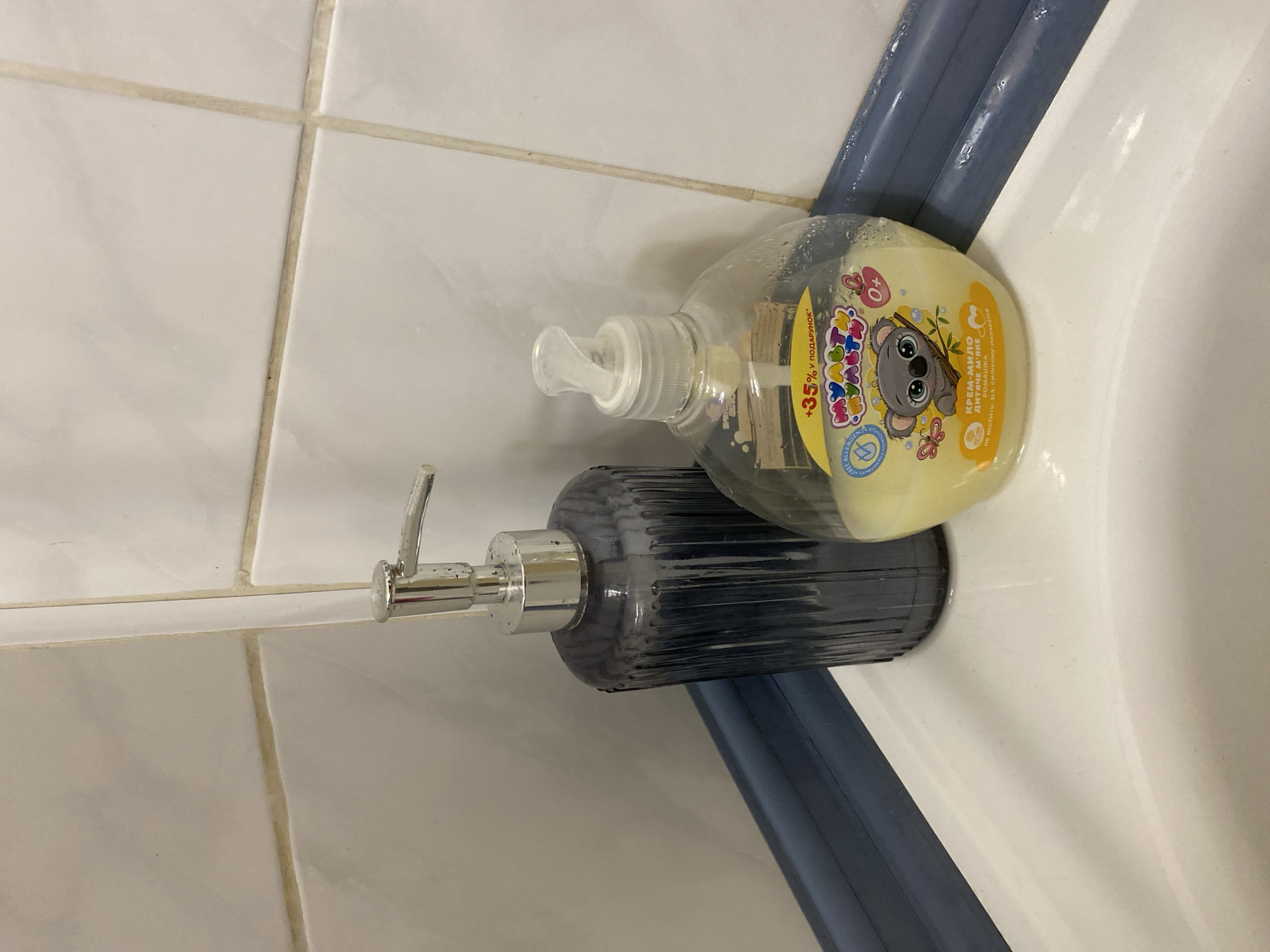 The image shows two soap dispensers on the edge of a white sink, likely in a bathroom. One dispenser is made of dark gray, ribbed glass with a silver pump mechanism. The other is a clear plastic bottle with a white pump, containing yellow liquid and decorated with a cartoon koala. The background consists of white tiled walls with light brown grout. A dark blue plastic trim runs along the bottom edge of the tiles. The scene is clean and well-lit, suggesting an indoor domestic setting. The location is not specified, so it cannot be definitively placed in Kryvyi Rih, Ukraine.