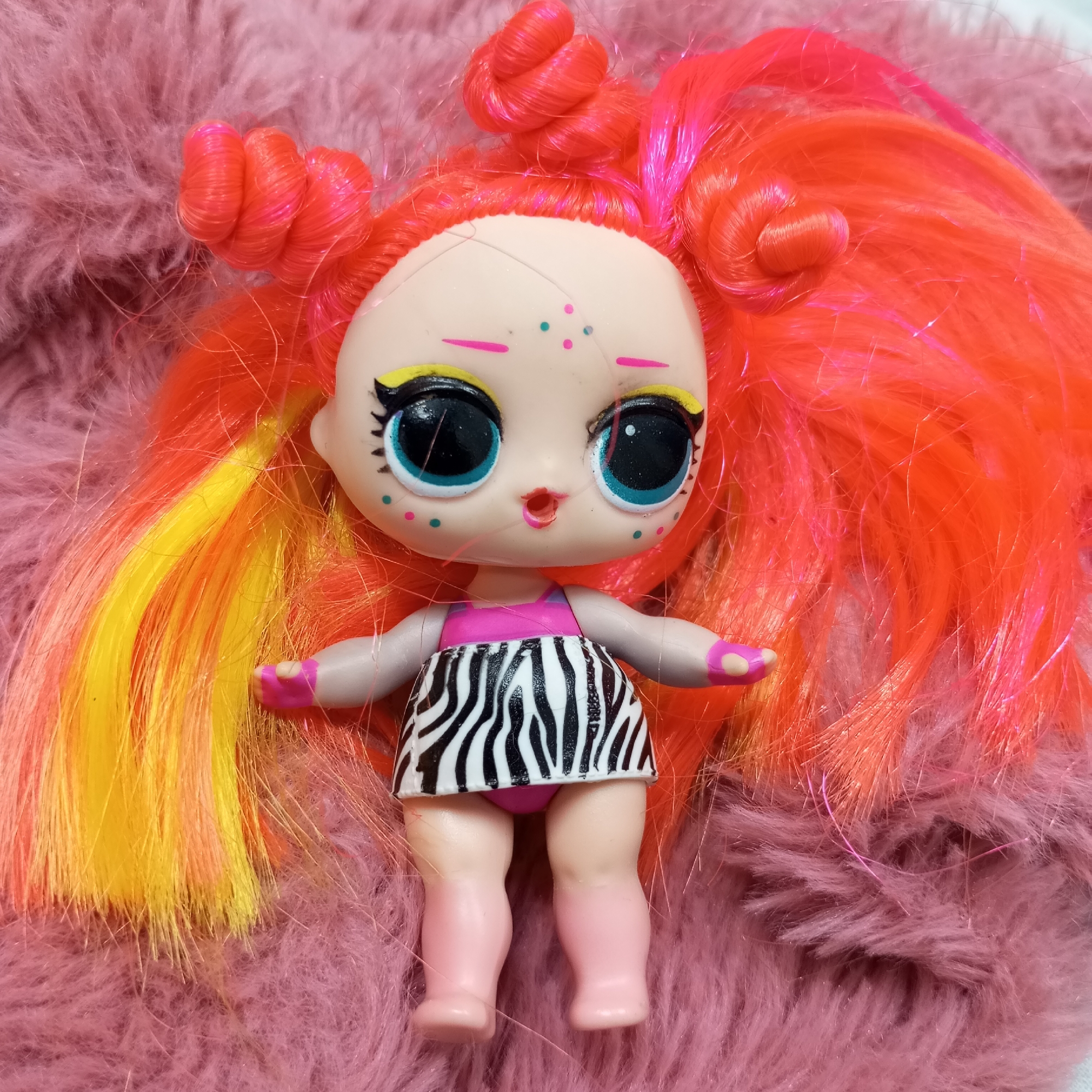 A top-down shot features a doll with vibrant, multicolored hair lying on a plush, pink faux fur surface. The doll has a light skin tone, large, round eyes with blue irises, prominent black pupils, black eyeliner, and bright yellow eyeshadow. Its face is detailed with small pink and teal dots resembling freckles on the cheeks and chin, and tiny pink lines with central dots on the forehead. The doll's lips are painted pink and slightly parted. Its hair is predominantly orange and pink, with striking yellow streaks, styled into several small, curly buns on top of its head. The doll is dressed in a black and white zebra-print skirt, underneath which pink molded underwear is visible. Its hands and molded feet are light pink, with pink accents on the hands suggesting gloves or painted nails. The soft, textured pink background suggests an indoor, cozy setting. No text is visible in the image.