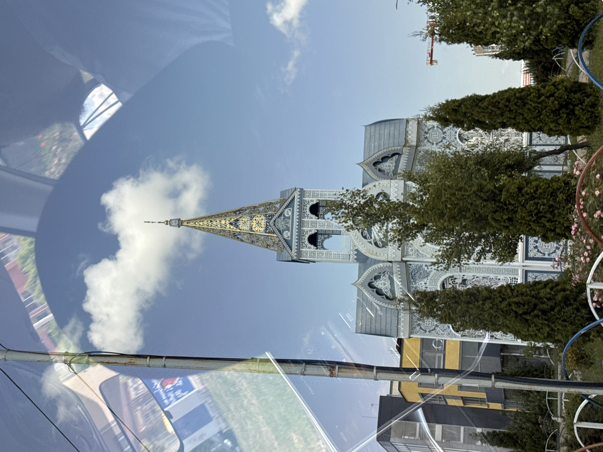 A unique, ornate building with a tall, gilded spire stands out against a clear blue sky with scattered white clouds. The building features intricate white and blue decorative patterns on its façade and multiple arched windows. It is partially obscured by lush green trees in the foreground, which include several tall, slender cypress trees. In the background, modern buildings, including a tall yellow and dark grey structure, are visible, along with a construction crane. The image appears to be taken from inside a vehicle, as evidenced by reflections on a glass surface in the foreground. This building is the Church of the Assumption of the Most Holy Mother of God in Mikhaylovsk, Russia.