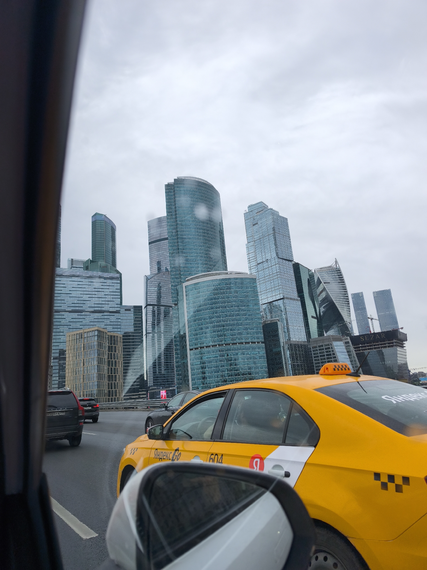 The image depicts a view from inside a vehicle, likely a car, on a wet road, with a cityscape in the background dominated by modern skyscrapers. The setting is likely Moscow, Russia, specifically the Moscow International Business Center (Moscow City) due to the distinctive architecture of the skyscrapers. A yellow taxi cab, marked with "Yandex Go" branding in Cyrillic script, is visible in the adjacent lane. Other vehicles, including an SUV, are also present on the road. The sky is overcast, suggesting a cloudy or rainy day. The image is taken through a car window, and a side mirror of the vehicle is visible in the foreground. The composition emphasizes the urban environment and the movement of traffic.