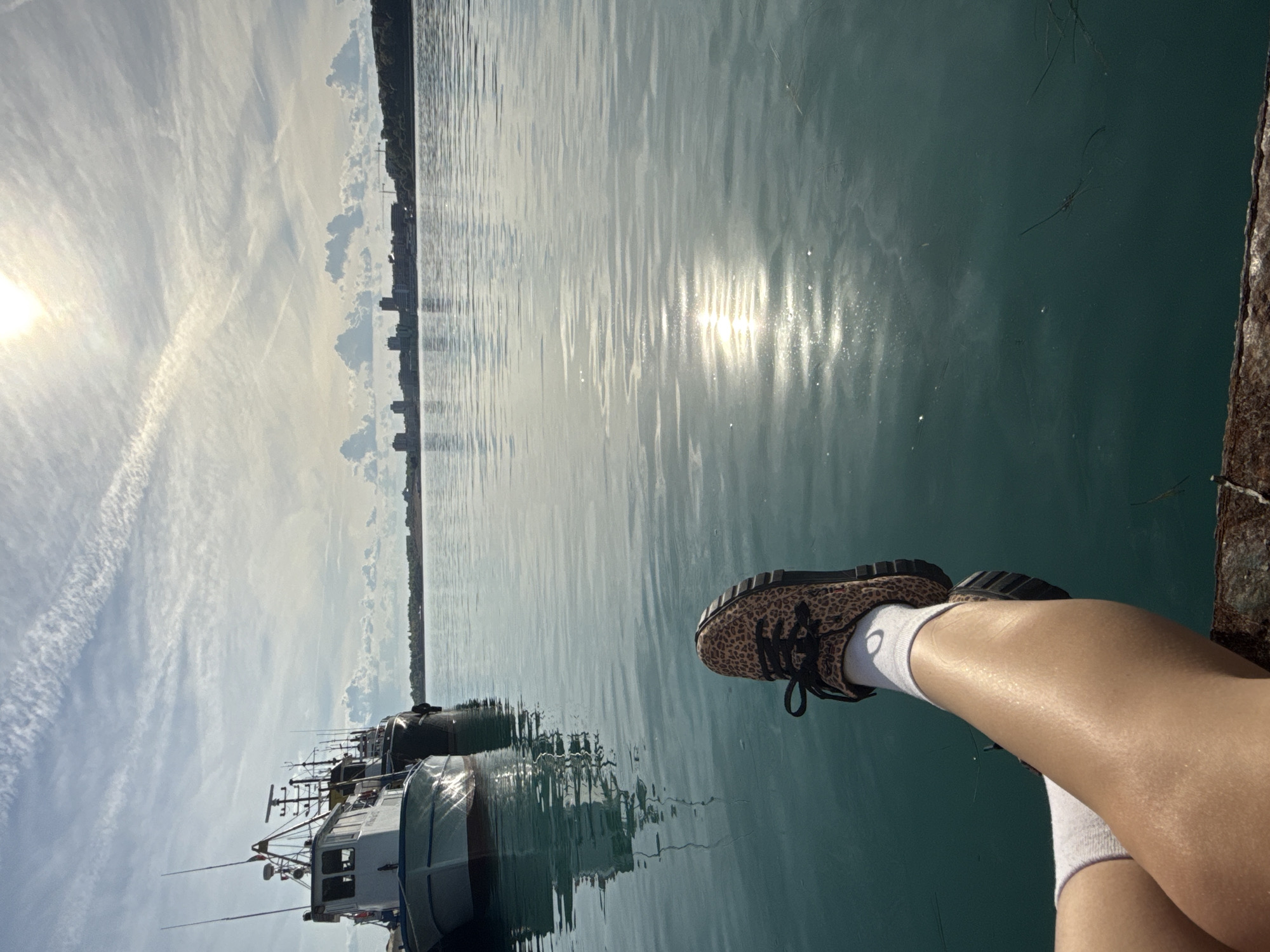 A horizontal, medium-shot captures a person's legs in white socks and leopard print sneakers dangling over a pier or dock. The person is looking out onto a body of water, likely the Black Sea, in Nesebar, Bulgaria, as evidenced by the boats in the background and the skyline on the horizon. The water reflects sunlight, creating a shimmering effect. The sky is partly cloudy with the sun visible. The edge of the pier or dock is visible on the right side of the frame.