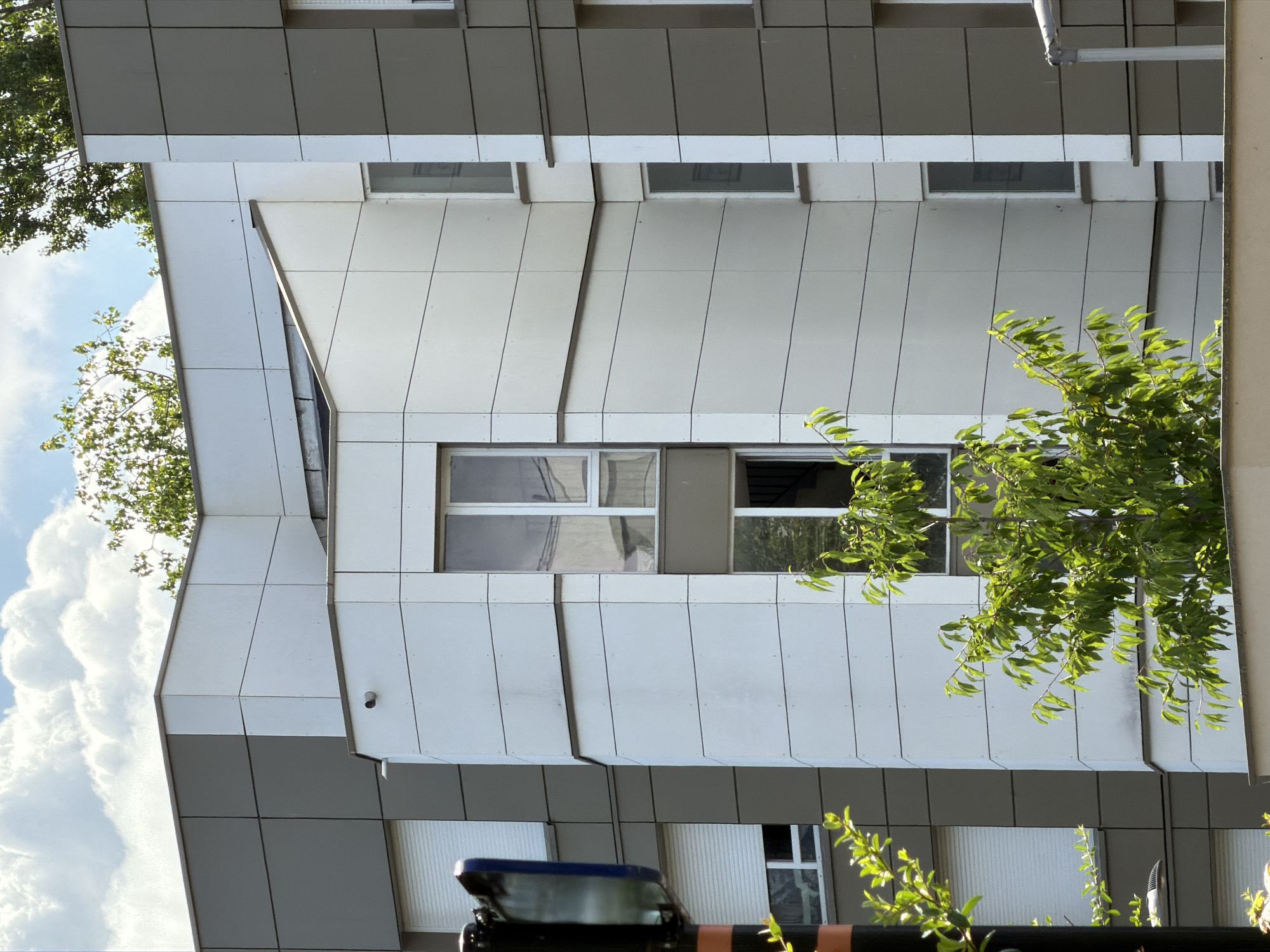 The image captures a segment of a modern building's facade, characterized by its angular design and metallic cladding. The building features several windows, some with visible reflections of trees and sky. Lush green foliage from nearby trees partially obscures the building, suggesting an outdoor setting with abundant vegetation. The sky appears bright with large, fluffy clouds, indicating daytime, likely sunny weather.
The composition is vertical, with the building occupying the majority of the frame. The architectural style is contemporary, with clean lines and a mix of light-colored panels that create a geometric pattern. One prominent window shows a clear reflection of the sky and clouds, while others reflect the surrounding greenery.
The overall impression is one of urban architecture set against a natural backdrop. The sunlight filtering through the leaves and reflecting off the building's surfaces creates a dynamic play of light and shadow. No people are visible in the image. The scene appears peaceful and serene, with no specific activity taking place. The location context is identified as Évry-Courcouronnes, France. There is no visible text in the image.