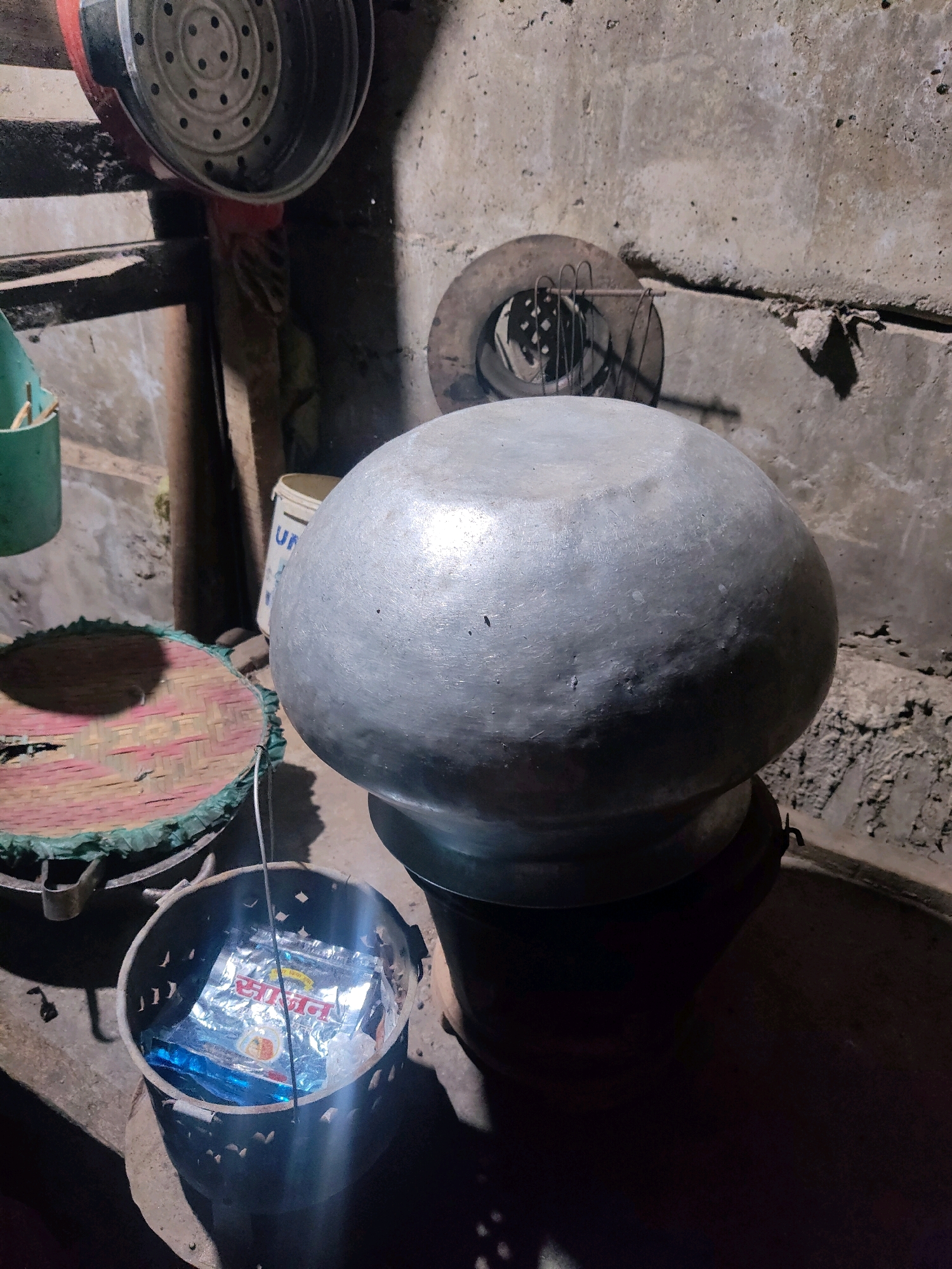 The image depicts a dimly lit indoor scene, possibly a kitchen or storage area, in Moreh, India. The focal point is a large, grey, pot-shaped object with a rounded top, resting on a dark base. To the left of the large pot, a round, woven mat with green edging is visible. In the foreground, a small, perforated metal container holds a silver packet with blue and red writing. Behind the large pot, a white bucket with the letters "UN" printed on it stands against a rough, concrete wall. A circular metal object with a grid pattern and wire elements is mounted on the wall above and to the left of the large pot. The overall environment appears rustic and somewhat cluttered.