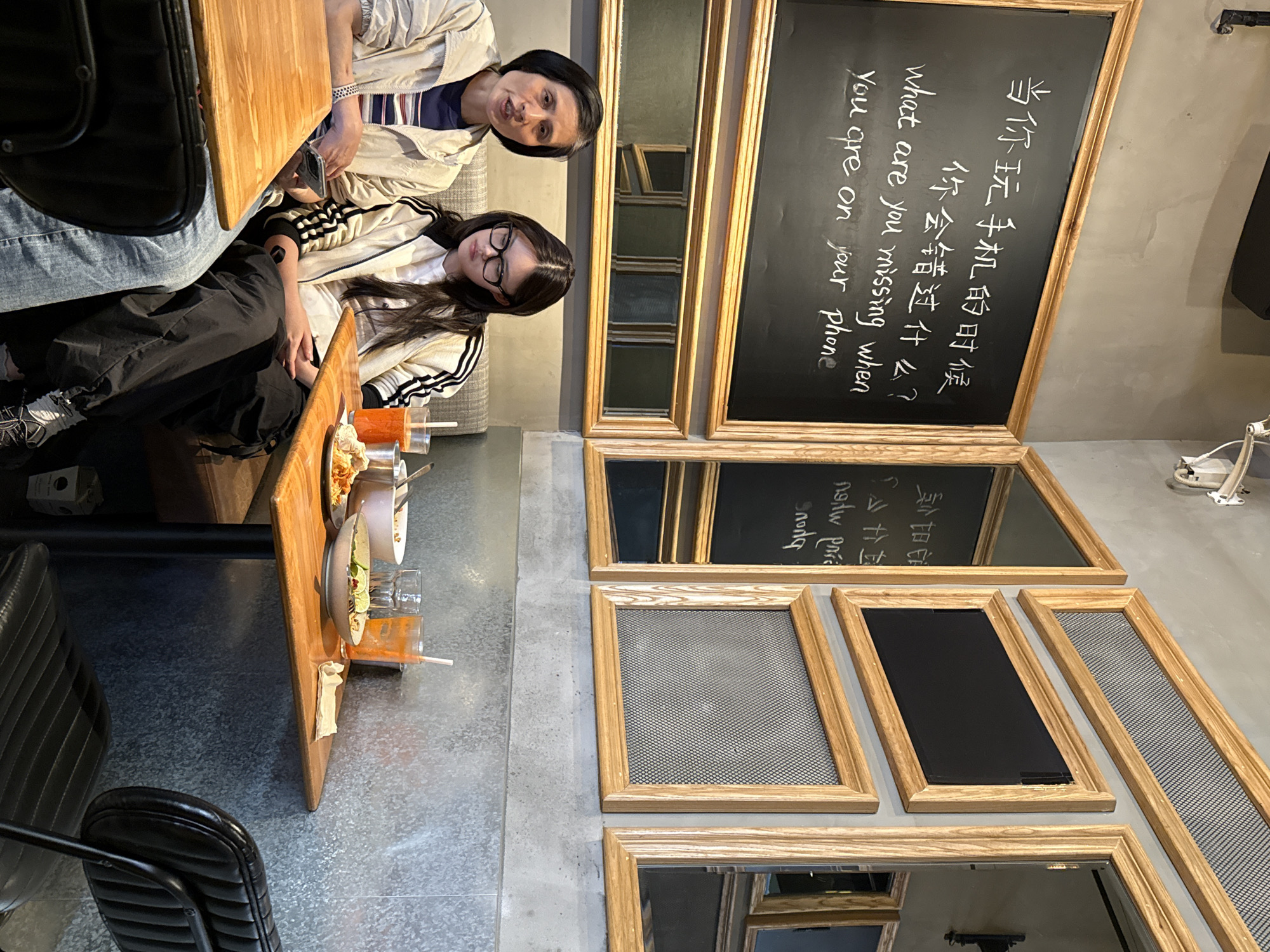This media file captures an indoor scene, likely a cafe or restaurant, in Yuzhong District, China. The image is taken from a slightly elevated, rotated perspective, showing two women seated at a wooden table on the left side.
One woman, with dark, short hair and wearing a light-colored jacket over a striped top, is looking directly towards the camera with a neutral expression. Beside her, another woman with long, dark hair and glasses wears a similar light-colored jacket with black stripes on the sleeves; she is looking down or away from the camera. The women are seated on a gray upholstered sofa.
On the wooden table in front of them, there are bowls of food and glasses containing orange drinks with straws, suggesting they are dining or having refreshments. Black chairs with ribbed backrests are also visible at other tables.
The background features a gray, concrete-like wall adorned with multiple framed items. Some frames contain mirrors, reflecting parts of the room, while others have a mesh or grid pattern. A prominent feature is a large blackboard within a wooden frame, displaying a message in both Chinese and English: "当你玩手机的时候 你会错过什么? what are you missing when You are on your phone". This message appears to be a prompt encouraging presence and discouraging excessive phone use, creating a reflective atmosphere in the setting. The overall aesthetic of the space is modern with industrial elements.