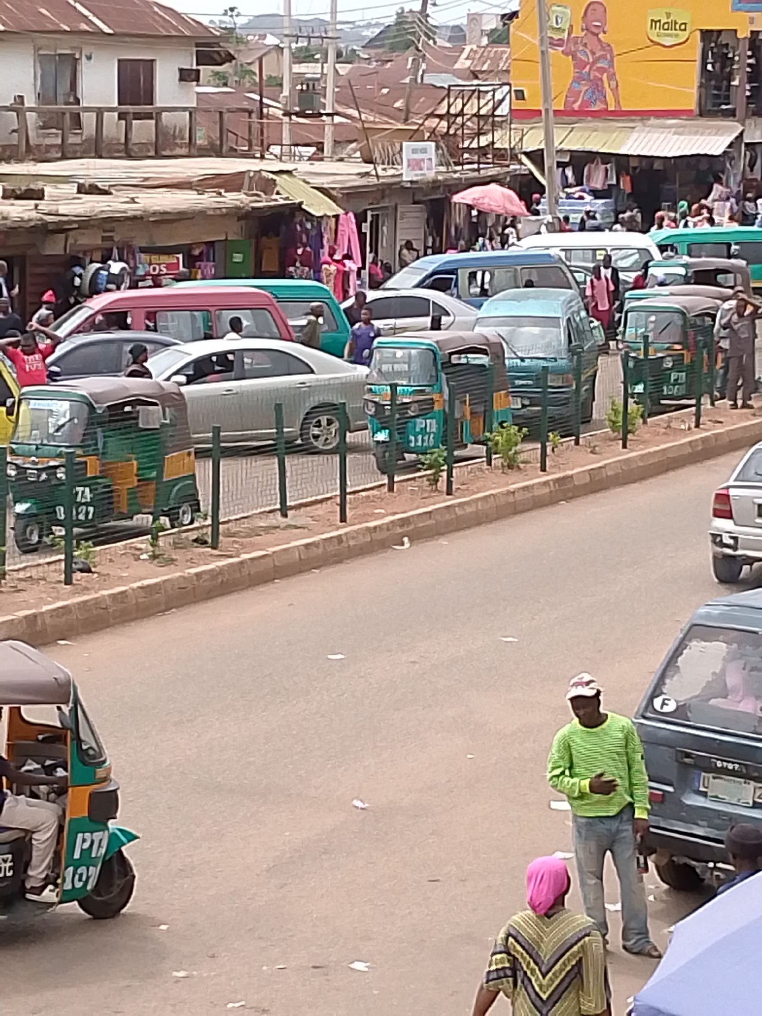 The image depicts a bustling urban street scene in an unidentified city. A row of green and yellow auto-rickshaws, some with license plates visible, is parked along the side of a road, separated from traffic by a low metal fence. Cars and vans fill the street, with people visible amongst the vehicles and on the sidewalks. Buildings line the street, including structures with metal roofs and awnings. A large billboard displaying an advertisement for "Malta" is prominently visible in the background. Pedestrians are present, including a man in a green shirt and jeans standing near a blue vehicle, and another individual with a pink head covering. The scene suggests a busy commercial area.