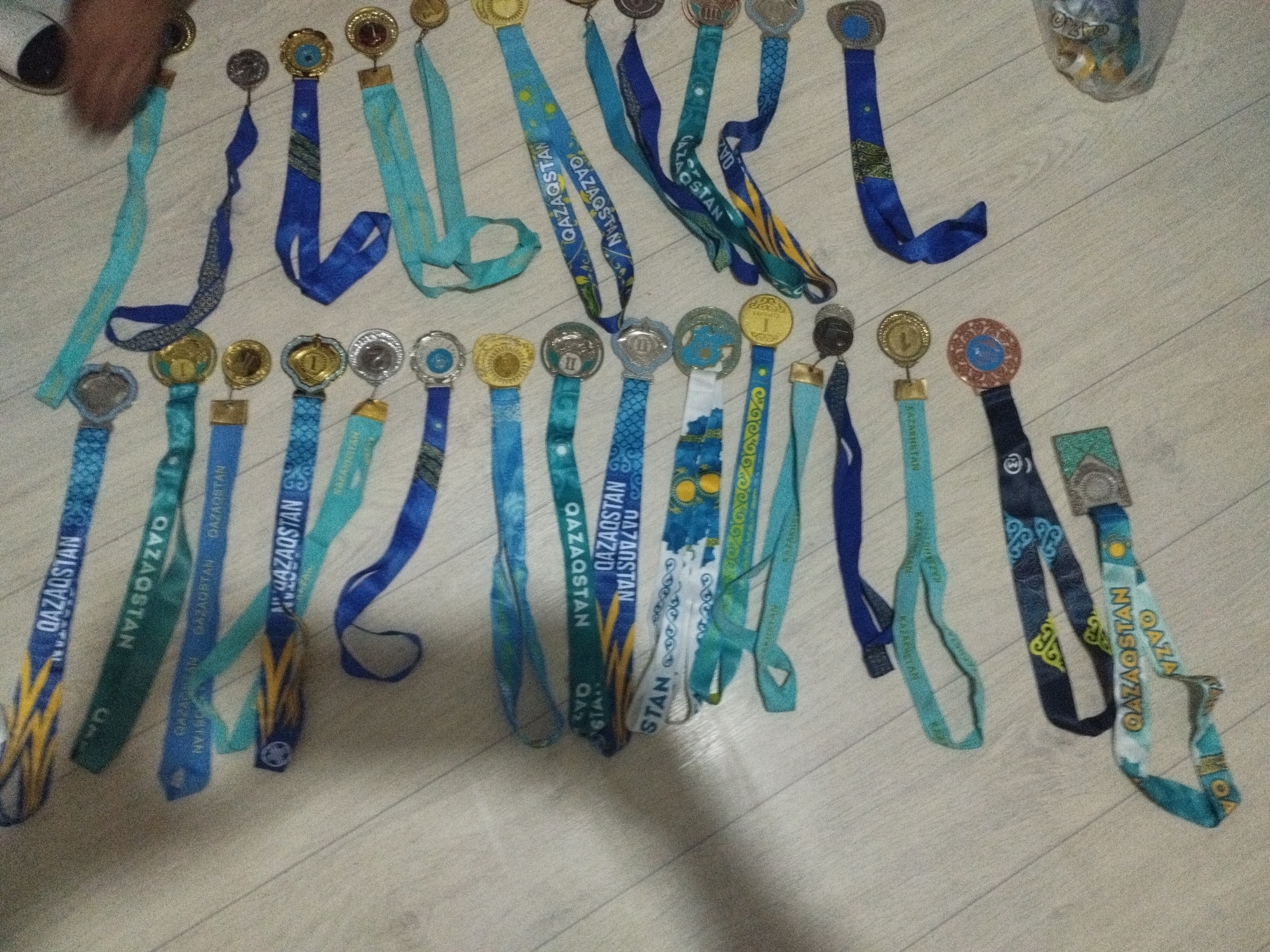 The image shows a collection of medals with ribbons, laid out on a light-colored wooden floor. The medals are of various designs and colors, predominantly featuring blue and gold. Many of the ribbons have text in a foreign language, possibly Kazakh or Russian, with "QAZAQSTAN" being visible on several. The medals appear to be awards or souvenirs. The setting is indoors, with a floor that resembles wood paneling. There are no clear indicators of the specific location within the city of Sarykol, Kazakhstan, beyond the presence of the text on the ribbons suggesting a connection to the country. A partial view of a person's hand holding one of the ribbons is visible in the upper left corner of the image.