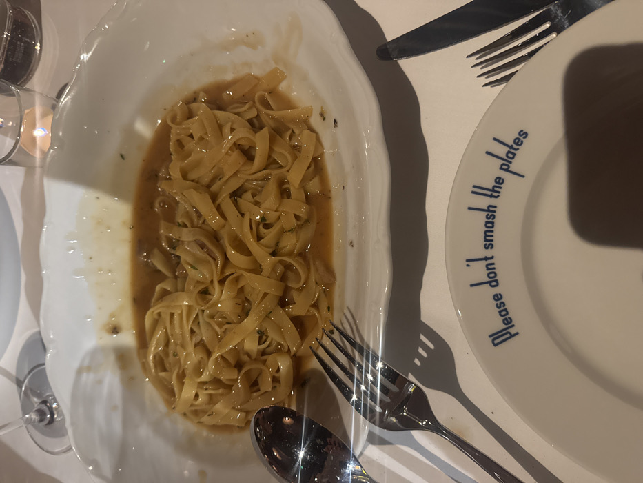 A high-angle photograph captures a dinner table scene within an establishment. A white, oval, scalloped serving dish holds a portion of tagliatelle-style pasta covered in a light brown sauce containing small particulate matter, possibly herbs or meat. The serving dish rests on a white tablecloth. To the left of the main dish, a silver serving spoon and a four-tine silver fork are present. In the upper left quadrant, a stemmed wine glass with clear liquid is partially visible. In the upper right, a glass tumbler containing a lit votive candle casts a warm glow. A white circular side plate is positioned at the bottom of the frame, displaying blue text along its rim that reads: "Please don't smash the plates". To the right of this side plate, a silver dinner knife and a four-tine silver fork are partially visible. Shadows are cast by the cutlery onto the white tablecloth. The location is Unknown, Unknown.