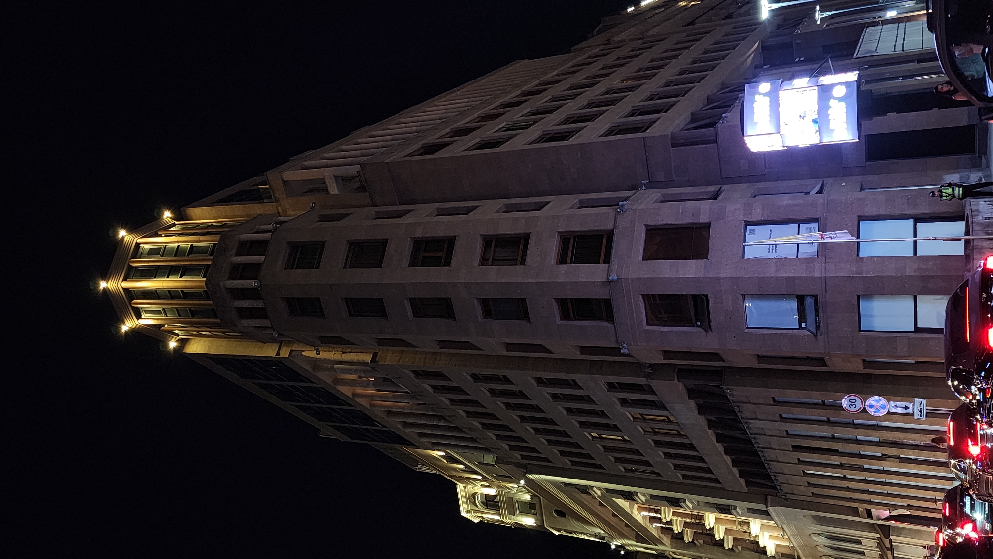 The image depicts the exterior of a large, multi-story building at night in Yerevan, Armenia. The building has a distinctive architectural style with classical elements and is illuminated by exterior lights, highlighting its facade and rooftop. On the left side of the building, a section with arched windows and ornate detailing is visible. The main part of the building is characterized by rows of windows and a prominent, angular corner. A digital billboard with illuminated panels is mounted on the right side of the building. In the foreground, several cars are stopped in traffic on a street, their headlights and taillights visible. Two individuals, one in a security uniform and another woman, are standing near the sidewalk. The scene is set against a dark night sky, suggesting an urban environment.