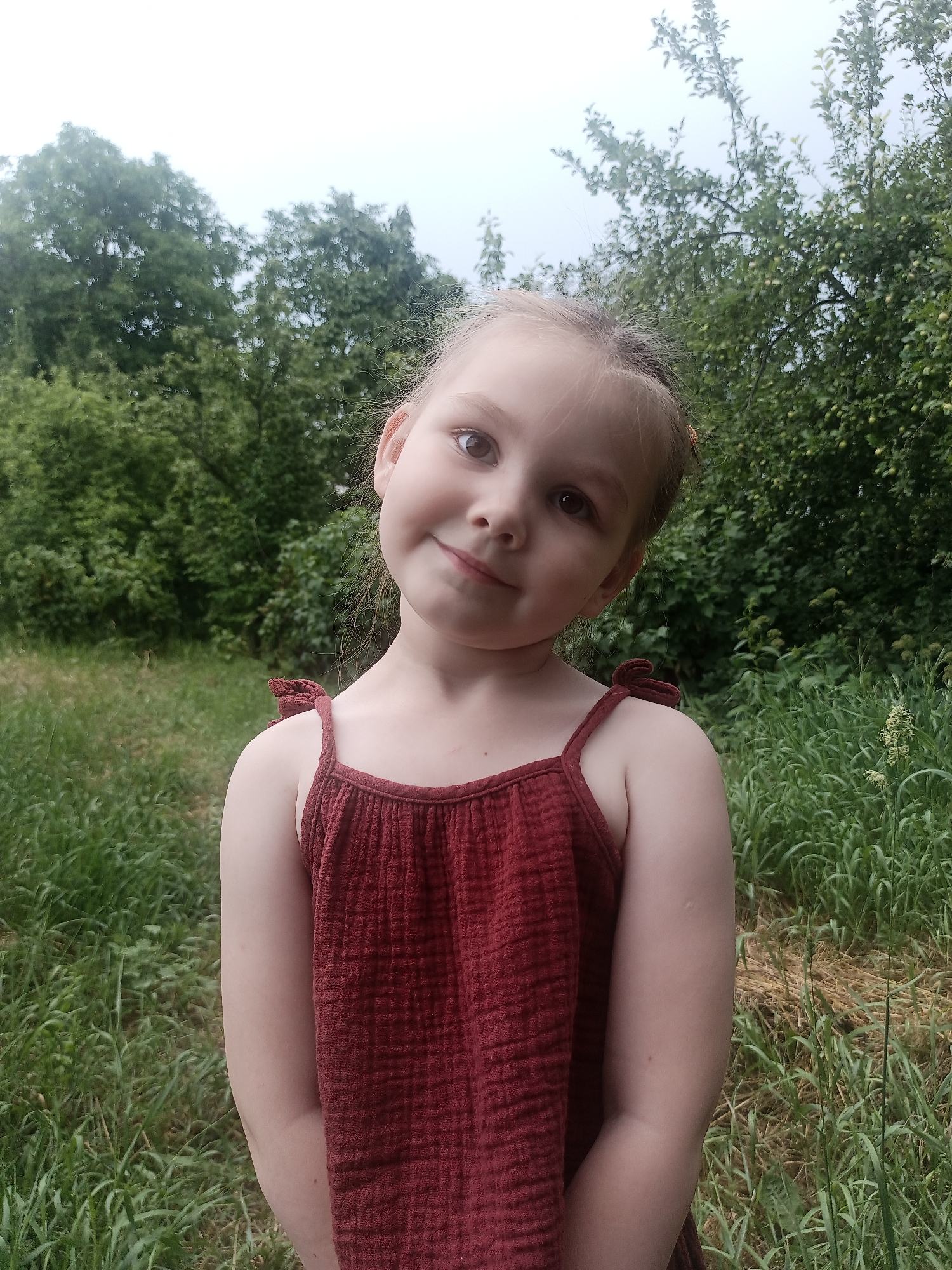 A young girl with blonde hair tied back and light brown eyes smiles at the camera. She is wearing a reddish-brown sleeveless dress with tie straps. She is outdoors in a natural setting, with lush green grass and trees behind her. The lighting suggests it is daytime, and the cloudy sky indicates possibly overcast weather. The girl appears happy and relaxed, and her head is tilted slightly. The background shows a dense arrangement of trees and tall grass, suggesting a rural or natural environment. There is no visible text in the image.