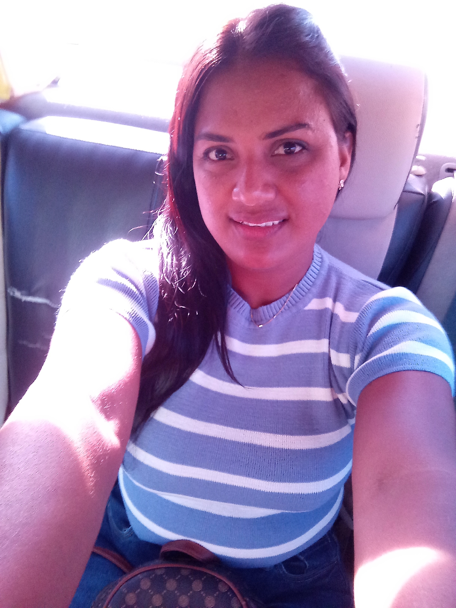 A woman is seated indoors, possibly in a vehicle given the headrest visible in the background. She is wearing a blue and white striped shirt and dark pants. A purse is resting on her lap. Her dark hair falls over her left shoulder. The location is not specified.