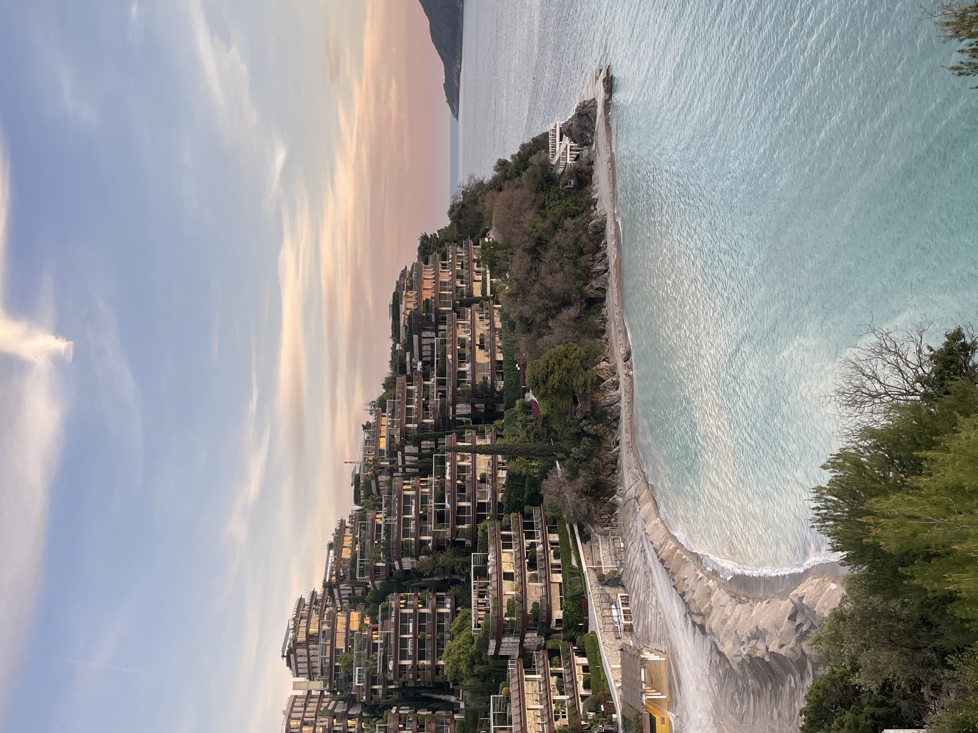 The image displays a coastal scene in Budva, Montenegro, specifically featuring the Dukley Gardens residential complex and its adjacent beach. The sky, occupying the upper third, shows a transition from pale blue at the zenith to soft orange and pink hues towards the horizon, indicative of twilight, with sparse, wispy clouds.
The midground is dominated by a steep, verdant hillside covered in dense vegetation, including numerous evergreen trees and shrubs. Integrated into this slope is a complex of modern, terraced residential buildings. These multi-story structures feature numerous balconies with glass railings and flat roofs, many of which are vegetated. The windows reflect the warm tones of the sky.
Below the hillside, a crescent-shaped beach of light-colored sand extends along the coastline. The sand appears groomed, exhibiting distinct parallel ridges near the water's edge. A small, thatched-roof structure and several white outdoor lounge furniture pieces are visible on the left side of the beach. A concrete or stone retaining wall separates the developed hillside from the sandy shore.
The sea, visible in the lower right and extending into the background, exhibits a clear, light turquoise coloration near the shore, transitioning to a deeper blue further out. Gentle ripples are present on the water's surface. In the distant background on the right, a dark, landmass or island protrudes from the sea, silhouetted against the horizon. No individuals or human activity are observed within the frame.